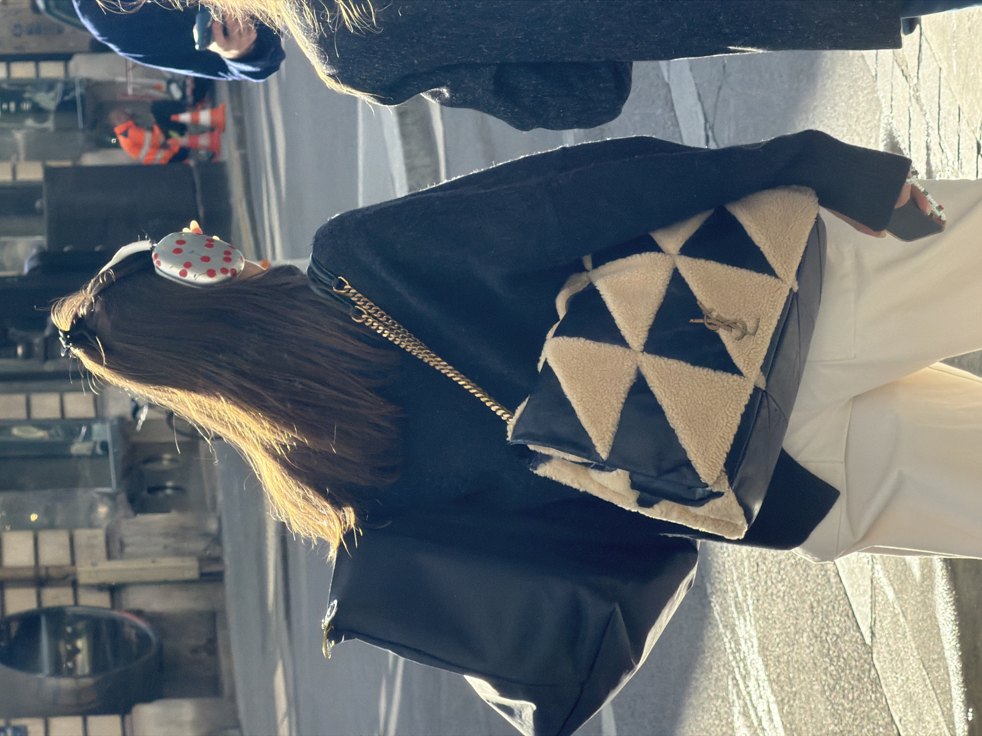 The image captures a person walking or standing outdoors in an urban setting, seen from the back and slightly to the side. The main subject has long, straight dark brown hair, illuminated by strong sunlight from behind, creating a golden halo effect. They are wearing a dark, possibly black, long-sleeved top or sweater, and white or light-colored trousers.
Key accessories include distinctive white headphones with red polka dots on the earcups, worn over the hair. The person is carrying two handbags: one is a plain black tote or shoulder bag with a silver-colored attachment visible near the top opening. The other, more prominent bag, is a designer shoulder bag with a gold chain strap. This bag features a geometric pattern of black leather triangles alternating with beige or cream-colored triangles made of a plush, furry material. A gold-colored YSL (Yves Saint Laurent) logo is visible on the front of this patterned bag. The person's left hand is visible near the patterned bag, holding a dark-colored phone or device with a beaded strap or charm.
The background is an out-of-focus urban environment, suggesting a street or sidewalk, consistent with the location context of Paris, France. Blurry structures, possibly buildings or storefronts, and a reflective surface are discernible. In the distant background, another person in an orange high-visibility jacket is faintly visible. The lighting indicates a sunny day, likely late afternoon or early morning, given the low angle of the sun casting long highlights and shadows. No other specific text is visible in the image.