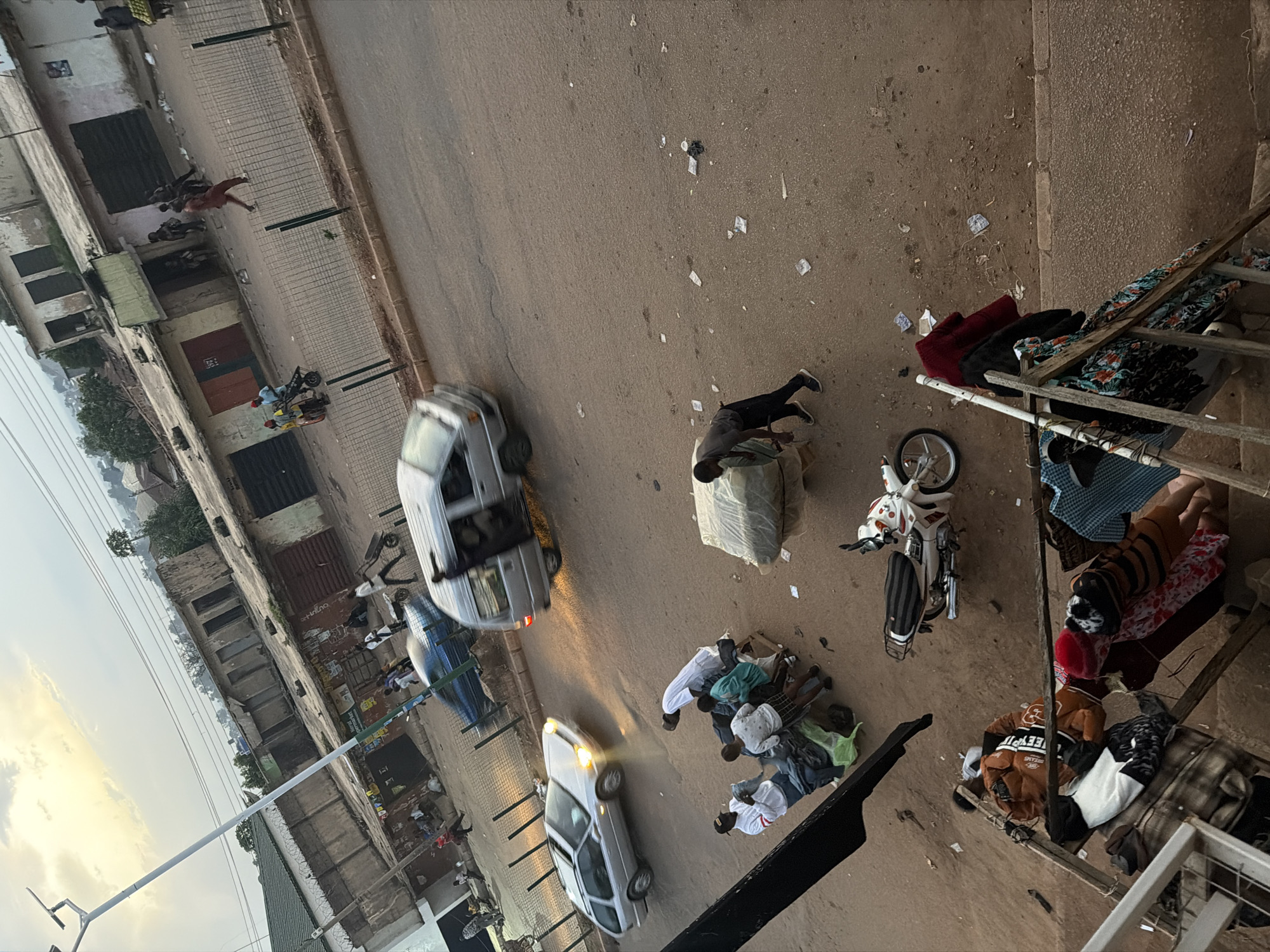 In the city of Bukuru, Nigeria, an outdoor scene unfolds. The sky above is overcast with hints of sunset. In the middle ground, a street is lined with buildings, some appearing weathered with closed metal shutters. A few people are visible on the street, some walking, others standing near shops. A silver van is in motion, its headlights on, and a silver station wagon is also moving. Several individuals are gathered around a wooden cart laden with goods. One person, wearing a dark shirt and black pants, is stooped over a large, wrapped bundle on the ground. Further down the street, a white motorcycle with black and white stripes is parked. In the foreground, a rack of clothing is displayed. Various items of clothing, including shirts and what appear to be blankets or large fabric pieces, are hanging or piled on the rack. The ground surface is predominantly dirt and asphalt, with scattered debris.