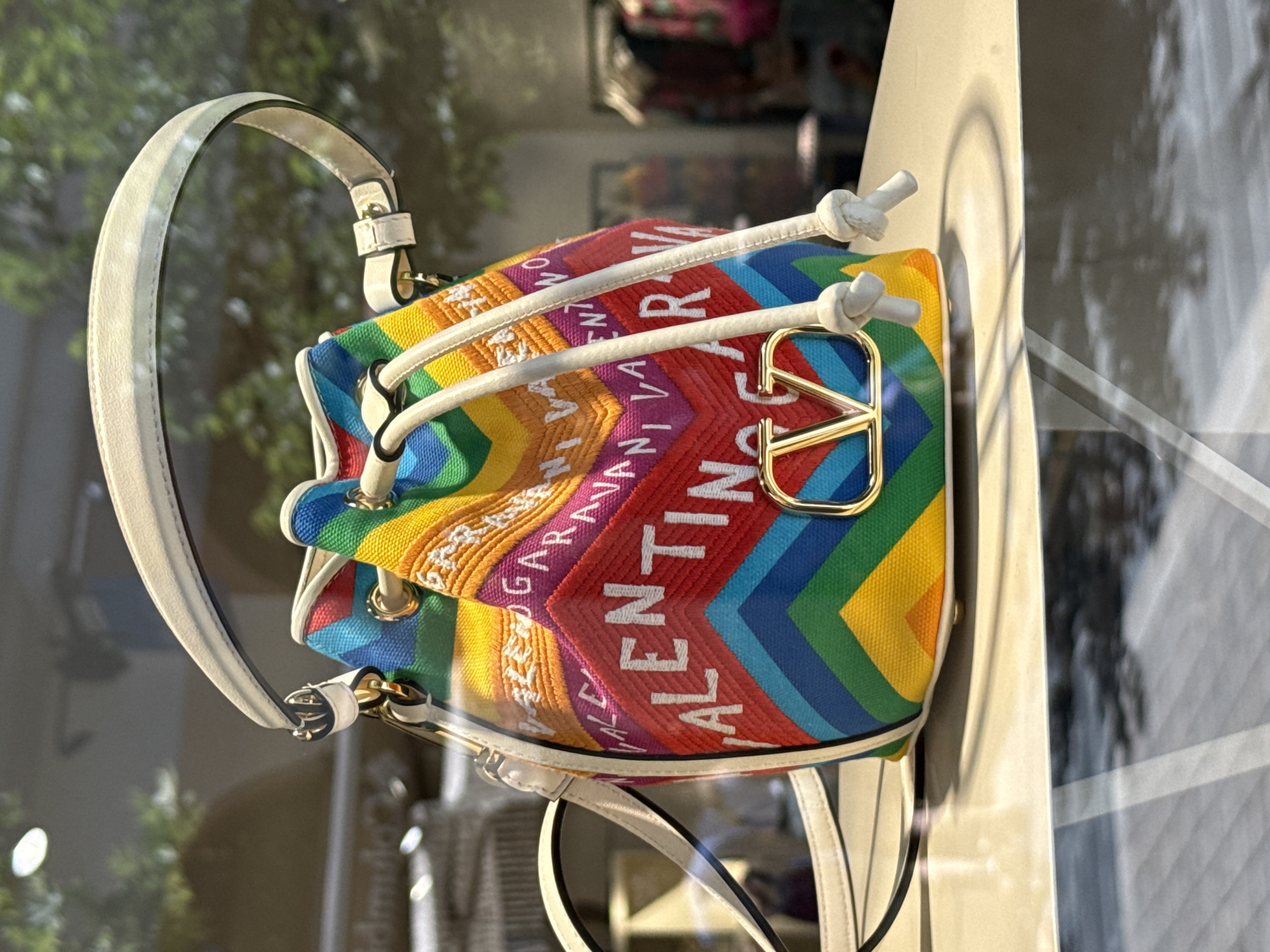 A vibrant, rainbow-colored bucket bag with white drawstrings and a cream-colored top handle is displayed in a store window. The bag is adorned with white lettering that reads "VALENTINO GARAVANI" in a repeating pattern. A prominent gold "V" logo is centered on the front of the bag. The bag is positioned on a light-colored display shelf. The background shows a blurred reflection of trees and possibly other store displays, suggesting an outdoor or indoor retail environment. The image is taken at eye level.
