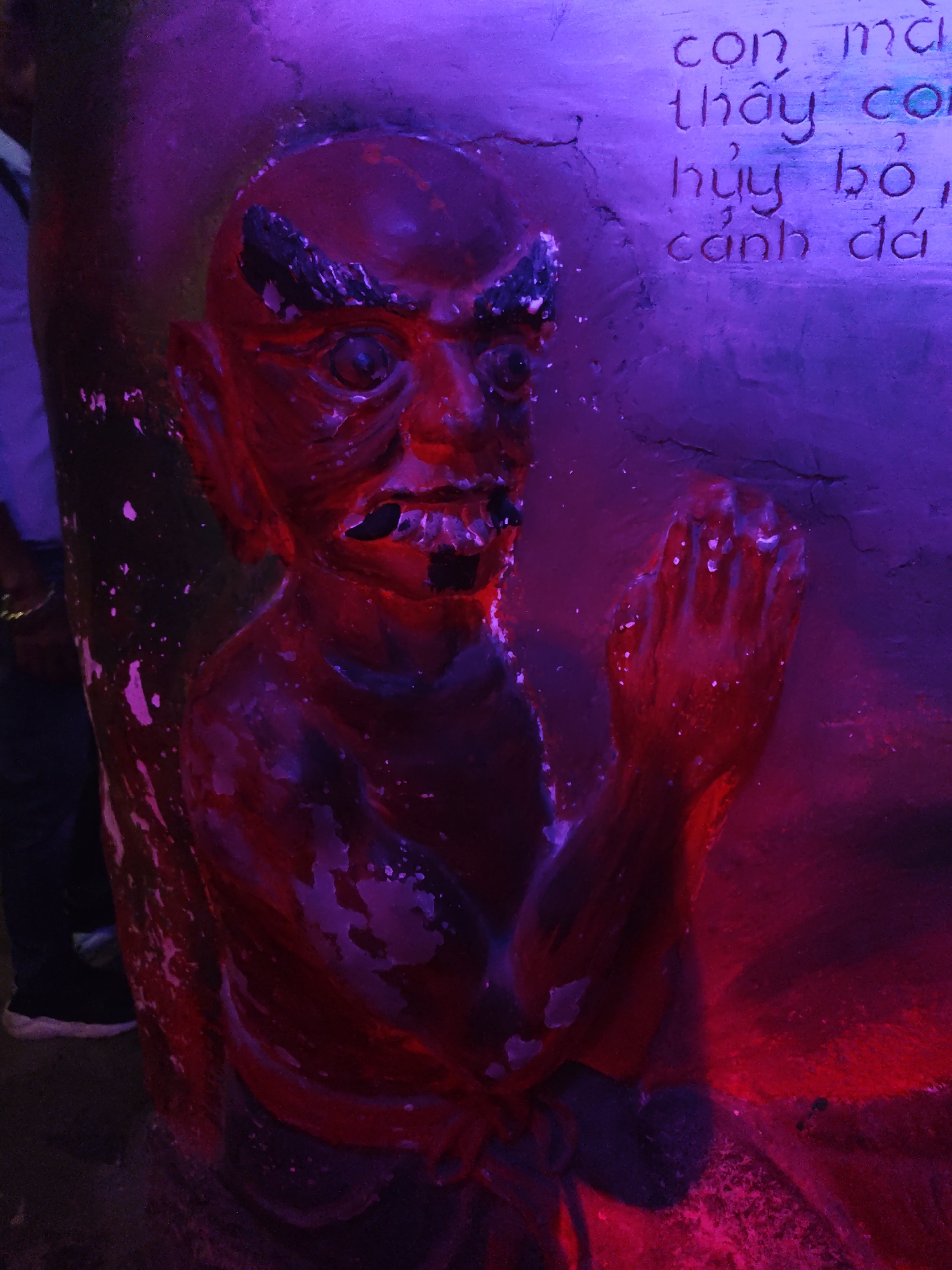 The image shows a close-up of a statue with a demon-like figure, illuminated by purple and red lights. The figure is red and appears to be made of stone or plaster, with its face contorted in an expression of distress or anger. It has large, dark eyes, a gaping mouth with dark openings, and prominent eyebrows. The figure's hands are clasped together in front of its chest, possibly in prayer or supplication. The statue is textured and appears to be weathered, with chipped paint or material in places, revealing lighter tones underneath. The overall effect is dramatic and somewhat unsettling, amplified by the intense lighting.
To the right of the statue, there is a textured surface with carved text. The text is in an East Asian script, likely Vietnamese, and appears to be carved into the stone. Some of the visible characters include "con ma", "thấy con", "hủy bỏ", and "cánh đá". These phrases suggest a narrative or inscription related to the statue, possibly a description or a cautionary tale. The background is dark and out of focus, with a hint of a person on the far left, wearing dark pants and white-soled shoes, suggesting the statue is part of an installation or a display in a public space. The lighting creates a moody atmosphere, with strong shadows and highlights, and the colors lend an otherworldly feel to the scene. No specific time of day or weather conditions are discernible from the image.