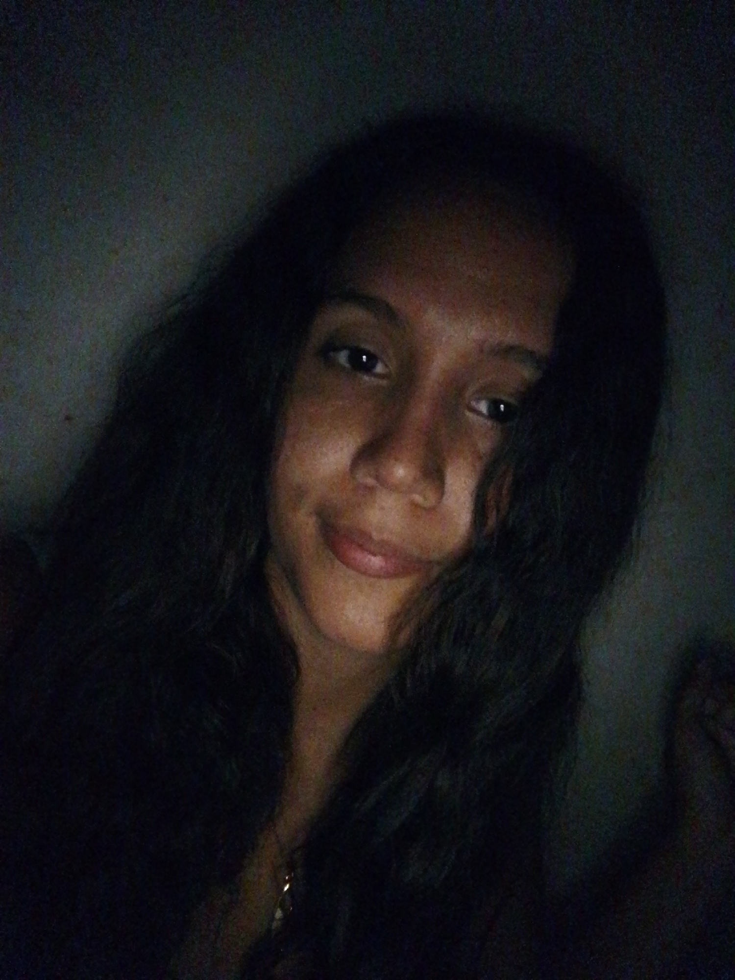 A medium shot of a young woman with long, dark, wavy hair. She is looking directly at the camera with a slight smile. The lighting is dim, and she appears to be indoors. The background is dark and indistinct. The location in Juan Griego, Venezuela, cannot be confirmed from the image.