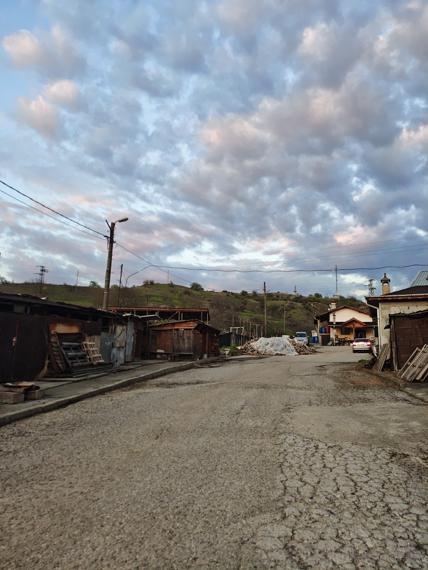 The image presents a street scene in a location that appears to be a somewhat rundown or impoverished area. The sky is filled with clouds tinged with blue and pink, suggesting either sunrise or sunset.
The road is cracked and shows signs of disrepair. On either side of the road are structures that appear to be garages or small sheds, many of which are constructed of corrugated metal or wood. Some are in a state of disrepair.
In the background, a grassy hill is visible. Power lines run across the sky.
Further down the street, a pile of firewood covered by a white tarp is present. There are two vehicles parked in the distance, one white and one red. A house or building with a light-colored facade is also visible.