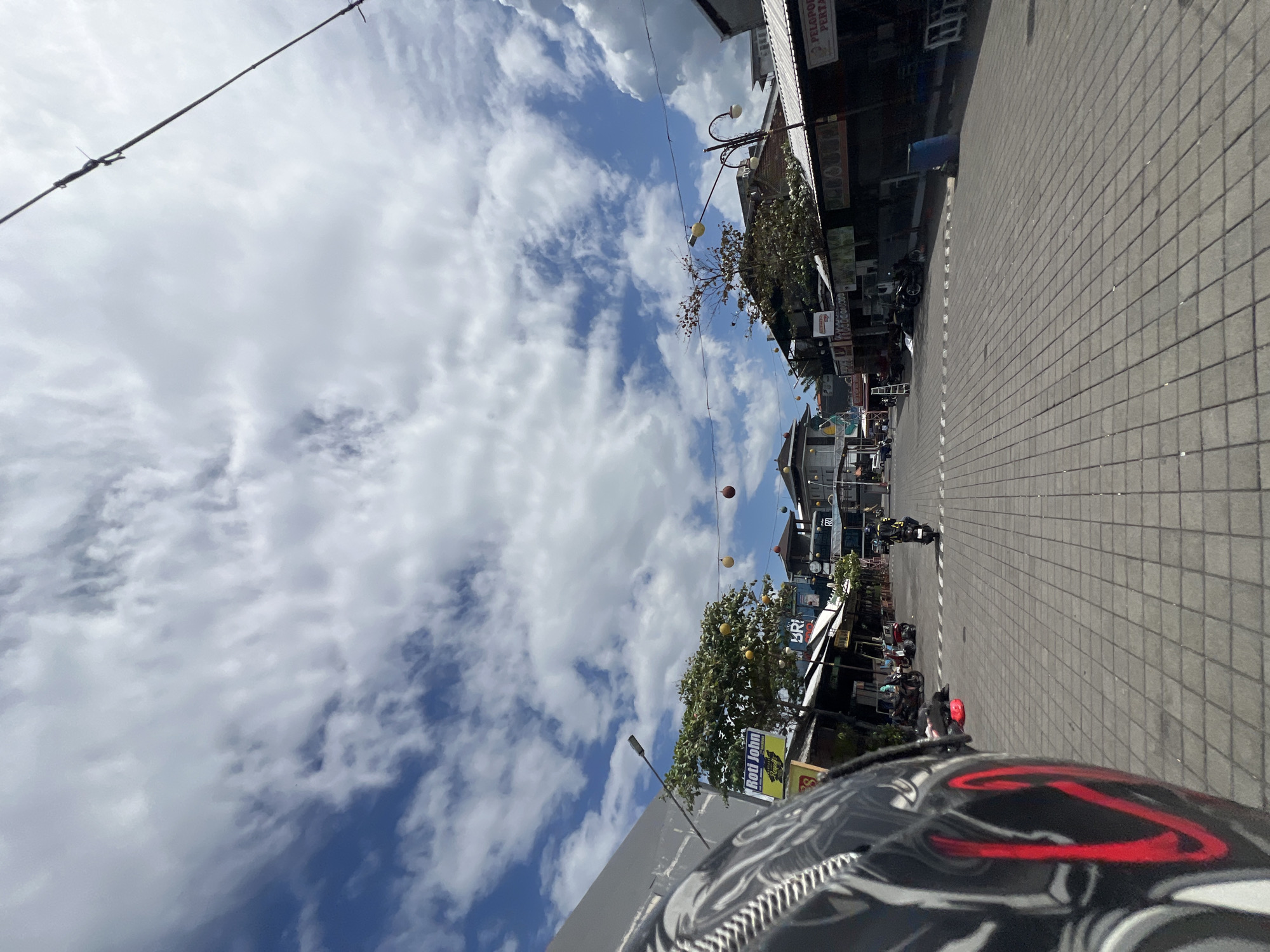 This image captures a street scene in Kuta, Indonesia, under a bright, cloudy sky. In the foreground, the left side of a black motorcycle helmet with red and white accents is visible. The street is paved with interlocking gray bricks, and it leads into the distance towards a cluster of buildings. On either side of the street are various shops and stalls with awnings. One prominent sign reads "Roti John." Several motorcycles are parked along the sides of the street, and a few people are visible in the distance, including one person riding a motorcycle down the street. Overhead, strings of colorful decorative lights are strung across the street. The overall impression is of a typical commercial street in a busy tourist area.