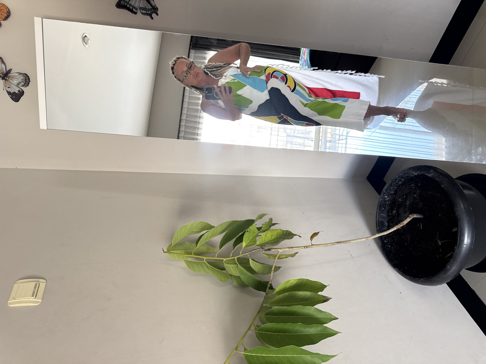 The image shows a woman's reflection in a mirror. The woman is wearing glasses, a dress with a colorful pattern, and has blonde braided hair. She is holding a smartphone and taking a selfie. In the foreground, there is a potted plant with long green leaves and a visible stem extending from the pot. The location is determined to be in Kuta, Indonesia, based on the setting and environment. The environment includes white walls, butterflies decorations and a black-and-white tiled floor.