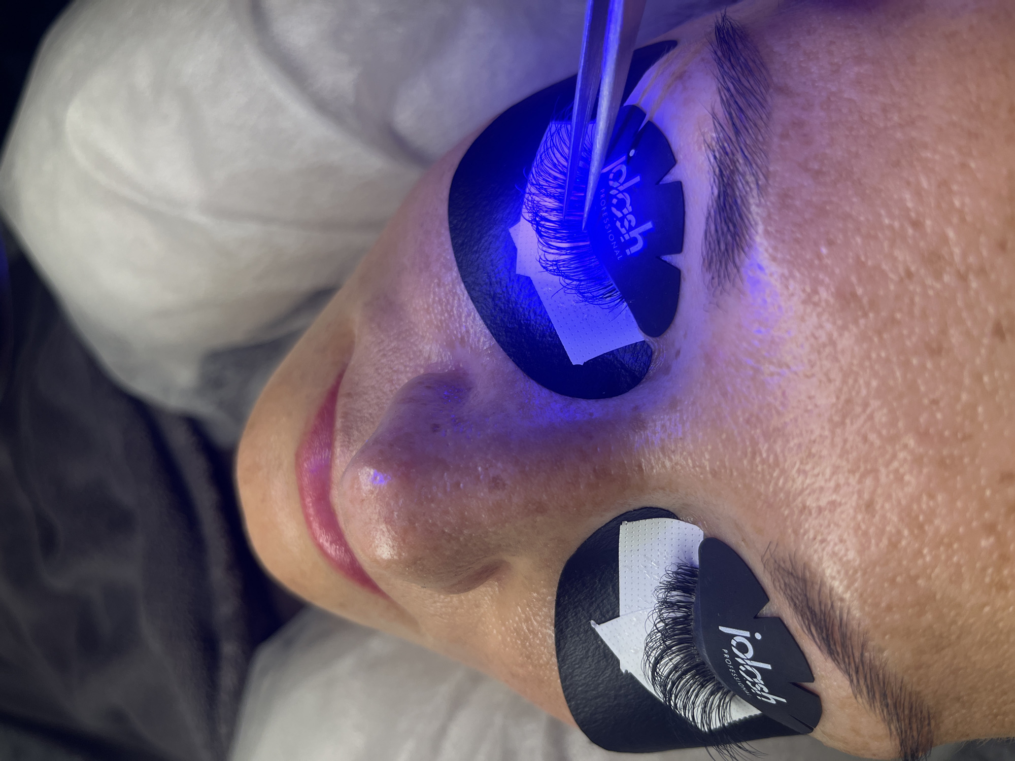A close-up, top-down view shows a person's face being prepped for eyelash extensions. The individual's eyes are closed, and under-eye pads with the "idash Professional" logo are in place. A blue light is being shone onto the eyelashes on the right eye, which are being manipulated with tweezers. The skin on the face shows some texture and pores. The background appears to be a soft, draped fabric, possibly a blanket or sheet. No specific location in Białystok, Poland is identifiable.