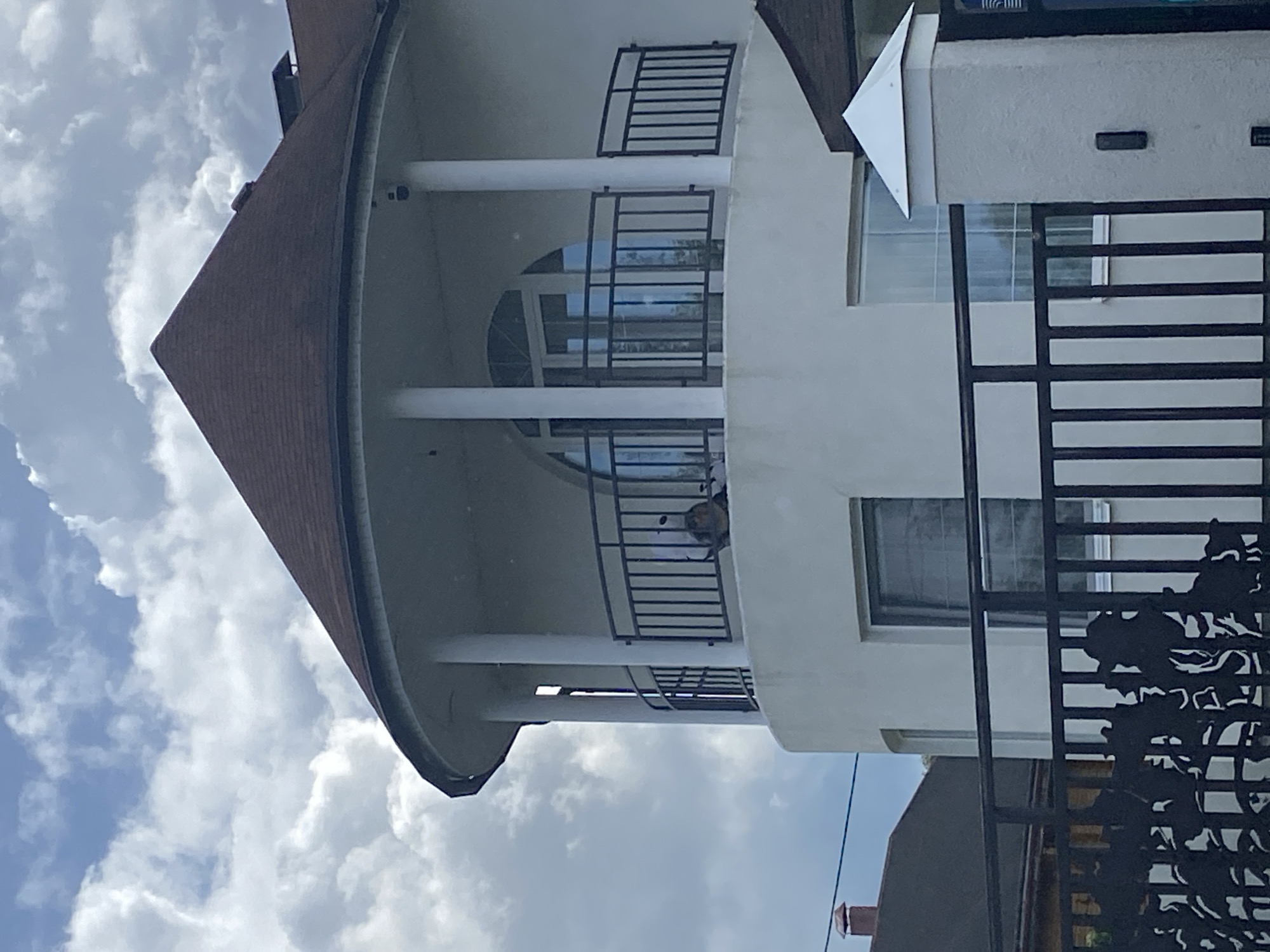The image captures a cloudy sky over a modern, two-story house with a rounded facade and a conical roof. The house is painted white, and features a balcony on the upper level with a black metal railing. A person is partially visible on the balcony, sitting down. Below the balcony, there are two large windows. A black metal fence with decorative elements is in the foreground, partially obscuring the view of the house and a portion of a neighboring building's roof. The scene appears to be set in a residential area of Votkinsk, Russia, judging by the architectural style and the presence of the ornate fence, which is common in some Russian regions.