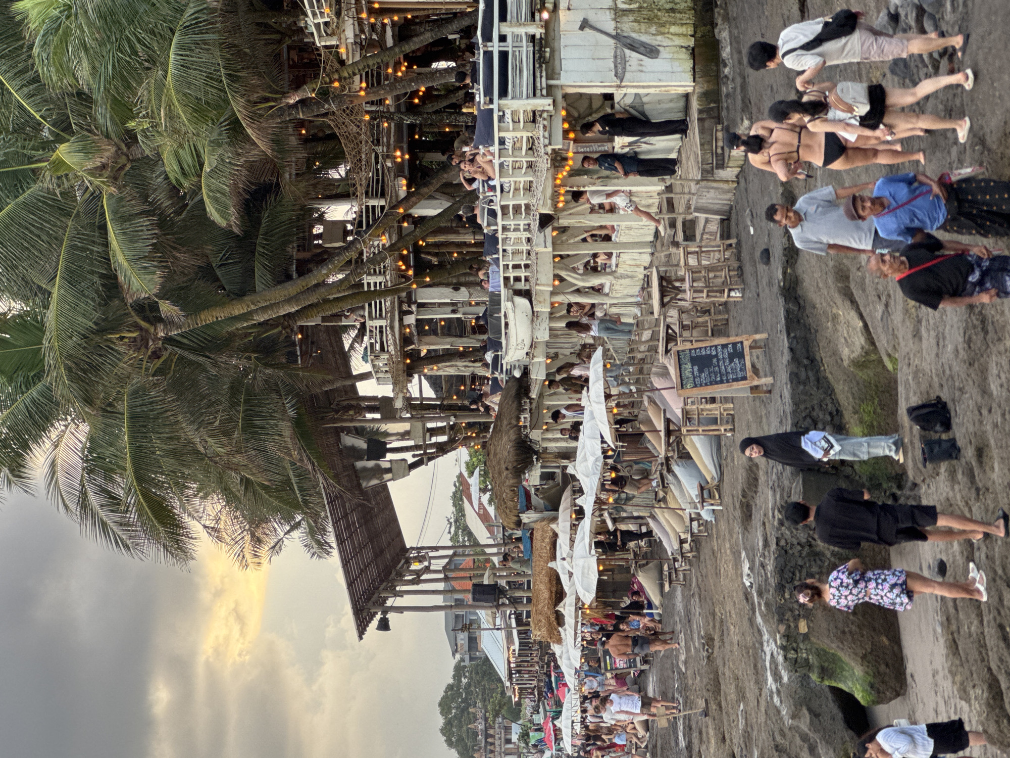 This media file captures a vibrant beachside scene in North Kuta, Indonesia, during what appears to be late afternoon or early evening, indicated by the warm, golden light filtering through a cloudy sky and the illuminated string lights.
The foreground showcases a bustling rocky and sandy beach area, where numerous people are present. Individuals are seen standing, walking, and gathering, dressed in a variety of casual attire including swimwear (bikinis, shorts), t-shirts, and dresses. One person is visible wearing a hijab and jeans.
To the left, large palm trees dominate the frame, their fronds contributing to the tropical ambiance. Interspersed within the landscape are multiple wooden structures resembling beach bars, cafes, or restaurants, built on various levels. These establishments are filled with people seated at wooden tables and benches, engaged in socializing, eating, and drinking. White parasols and umbrellas are positioned over some seating areas. Decorative string lights are strung between the structures and palm trees, creating a festive glow.
Notable details include a wooden sign with an oar, displaying partially visible text that appears to read "NO ENTRY." A portable chalkboard menu is also visible, listing various items with some illegible text and prices such as "30k." The overall impression is one of a lively and popular leisure destination, with people enjoying the tropical setting and social atmosphere.