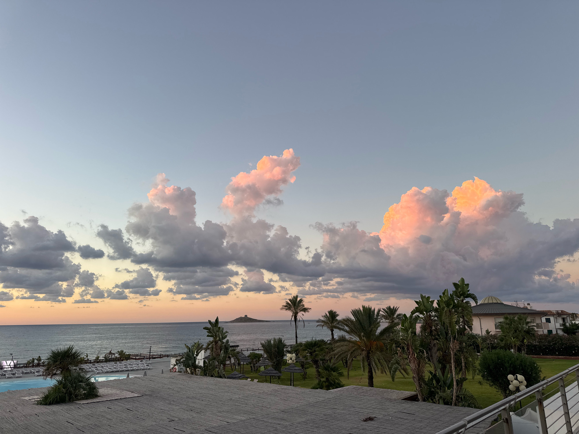 The image depicts a serene coastal scene at dusk, likely in Capaci, Italy, characterized by a tranquil sea meeting a softly lit sky. The horizon is painted with hues of orange and pink, transitioning into a pale blue overhead. Fluffy clouds, tinged with pink from the setting sun, are scattered across the sky.
In the foreground, a rooftop with a thatched covering is visible, suggesting a building or resort. Beyond this, a swimming pool with lounge chairs is partially seen, indicating a recreational area. Further back, a beachfront is lined with rows of empty lounge chairs and umbrellas, suggesting a quiet time of day. Palm trees and other lush greenery dot the landscape, adding to the tropical ambiance.
In the distance, a small island with a tower-like structure stands against the sea, a distinct landmark. The overall atmosphere is one of peacefulness and calm, with the natural beauty of the coast and the soft twilight colors creating a picturesque view. There are no people visible in the scene.