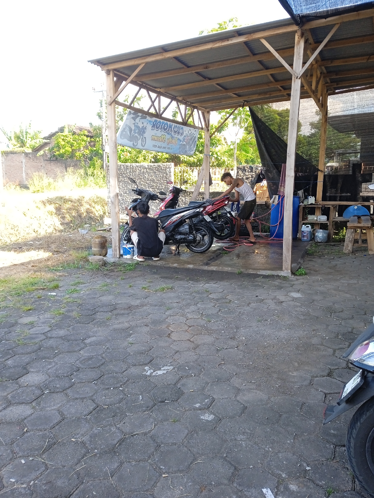 The image depicts an outdoor motorcycle washing station located in Cangkring, Indonesia. The station is sheltered by a wooden structure with a corrugated metal roof, partially covered with black netting. Two men are present: one is crouched near the front of a black and red motorcycle, appearing to be cleaning it with a brush and buckets of water. The other is standing and washing the back of another, similar motorcycle with a hose. A blue plastic barrel, various containers, and a wooden shelf unit holding bottles and cleaning supplies are visible near the washing area. The foreground shows a paved area composed of hexagonal concrete blocks. In the lower right corner, the front fender and wheel of another motorcycle are partially visible. In the background, there's a grassy area with a low concrete wall and trees. A sign advertising "Motorwash" is hung from the front of the shelter.