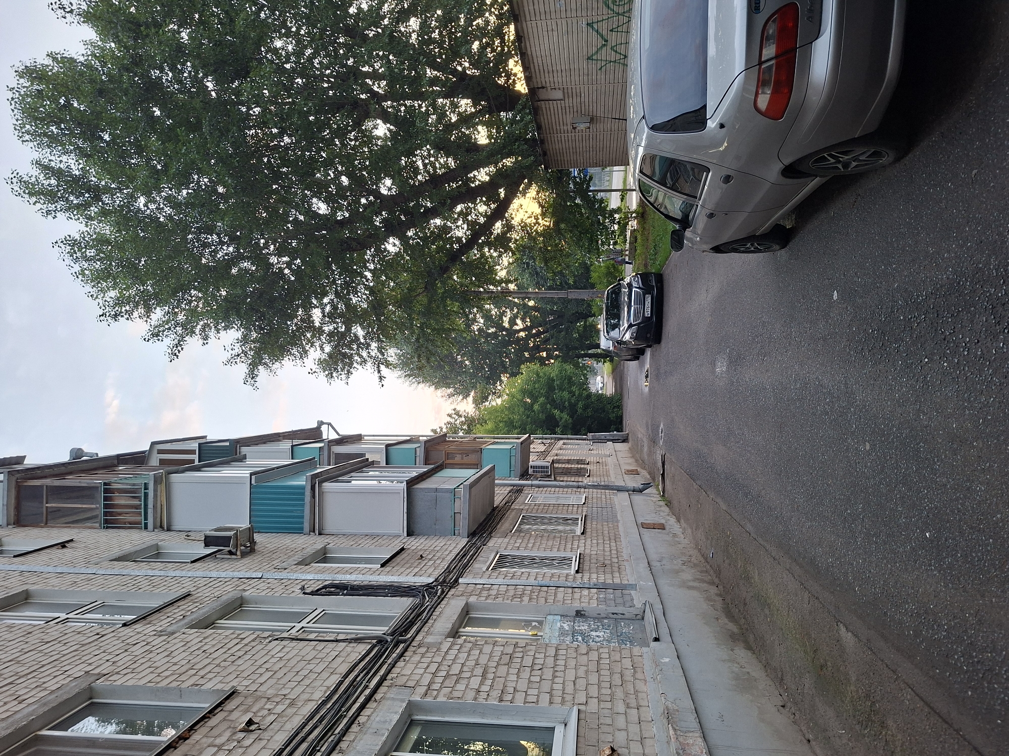 The image depicts a street scene in Kazan, Russia, on a cloudy day. On the left, a pale brick apartment building runs along the side of the frame, featuring multiple windows, balconies with teal accents, and external wiring. In the foreground, a gray sedan is parked on the right side of the asphalt road. Further down the road, two other cars, a dark sedan and a lighter colored sedan, are parked. Trees line the right side of the road behind a corrugated metal structure. The sky is overcast with a few patches of lighter sky visible towards the horizon.