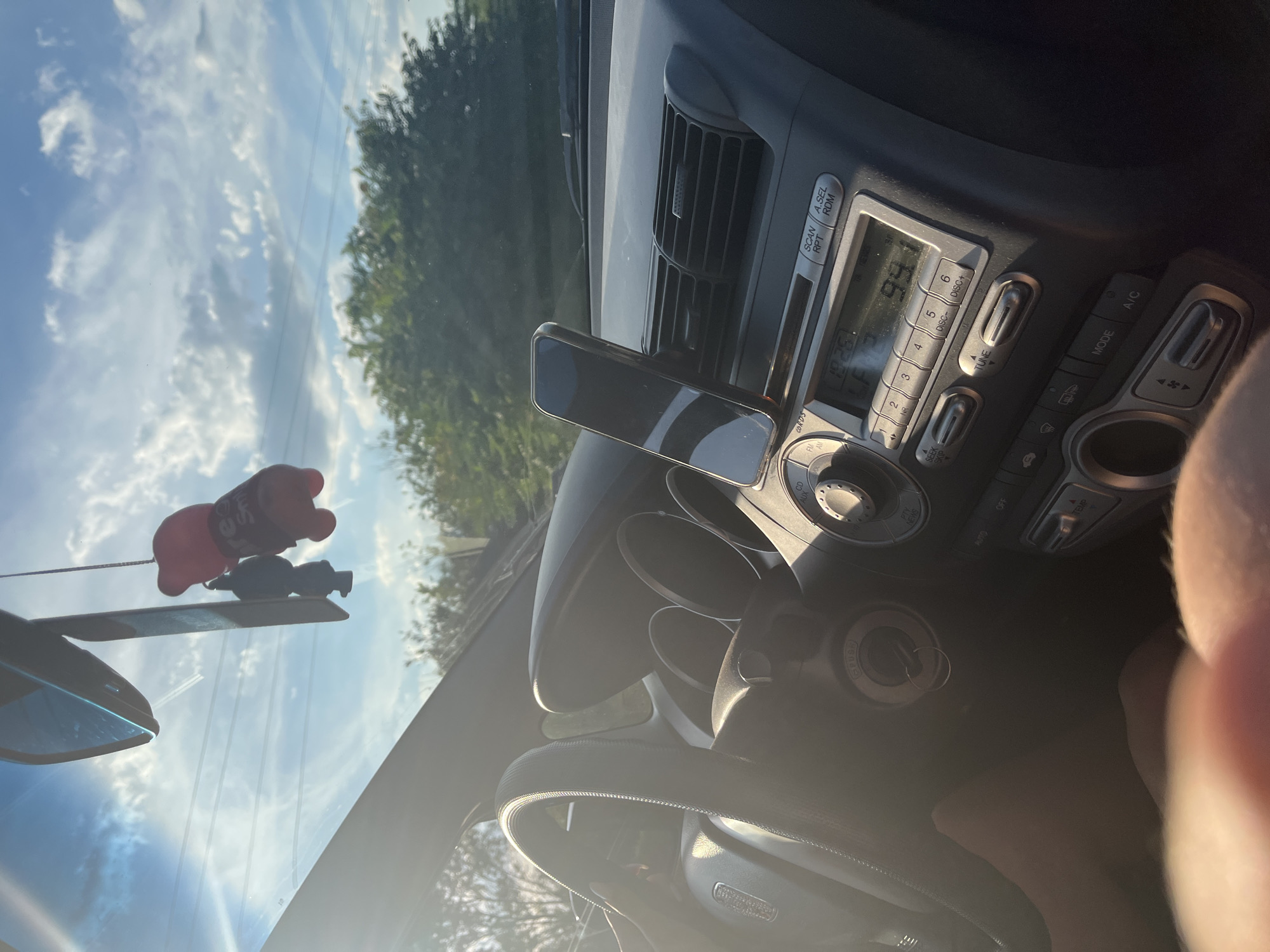 The image shows the interior of a car, looking towards the dashboard and windshield. A smartphone is mounted on a vent to the right of the steering wheel. The steering wheel itself is partially visible on the left, with a hand resting on it. The car's radio display shows "19:25" and "99.1". Outside the car, the sky is visible with clouds and sunlight, and trees can be seen. There are also two decorative air fresheners hanging from the rearview mirror. There is no indication that this image was taken in Dalgo Pole, Bulgaria.