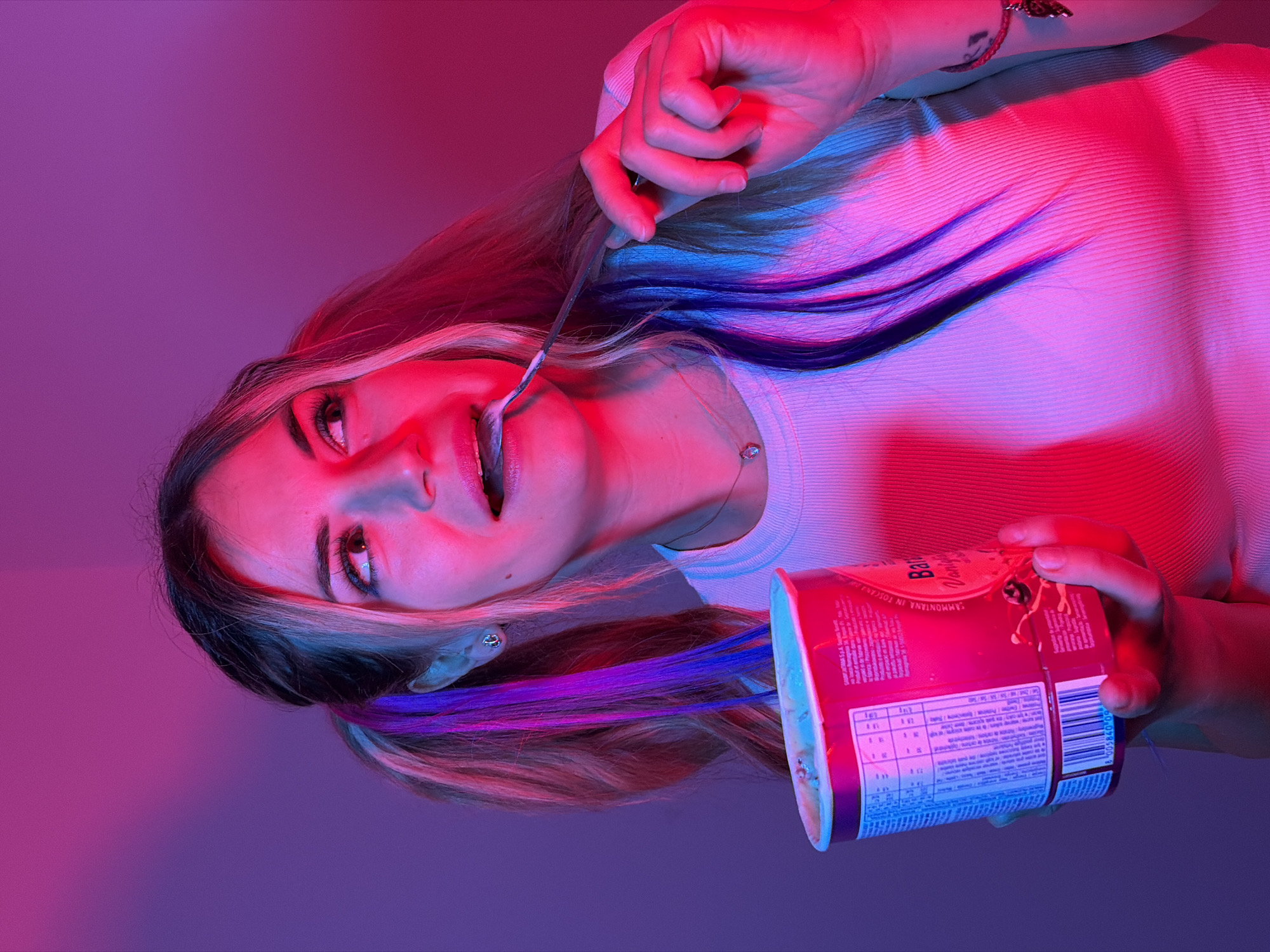 A woman with blonde hair, styled in two pigtails with streaks of blue and purple, is holding a tub of ice cream and eating it with a spoon. She is wearing a white ribbed tank top and has a small pendant necklace. The background is a gradient of purple and pink, illuminated by colored lights. The scene appears to be indoors, and no specific location in Budva, Montenegro is discernible from the image.
