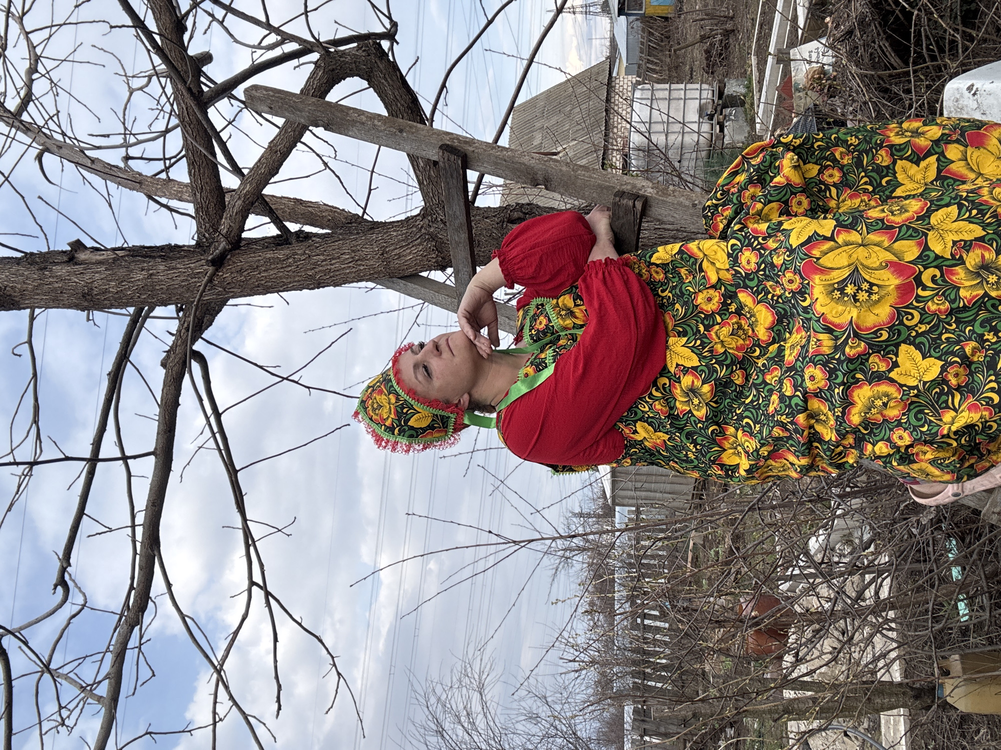 A woman in traditional Russian clothing sits on a wooden ladder propped against a tree in what appears to be a rural or semi-rural area. The woman is wearing a colorful floral dress and a matching headscarf. Her dress has a red bodice with green trim. She rests her head in her hand, appearing to be in a contemplative pose.
Behind the woman are bare trees and branches, suggesting it is winter or early spring. In the background, there are houses and utility lines, hinting at a residential area. The location is described as Chaikovsky, Russia, implying that the setting may be a home or garden in this city. The scene has a quiet, almost melancholy atmosphere.