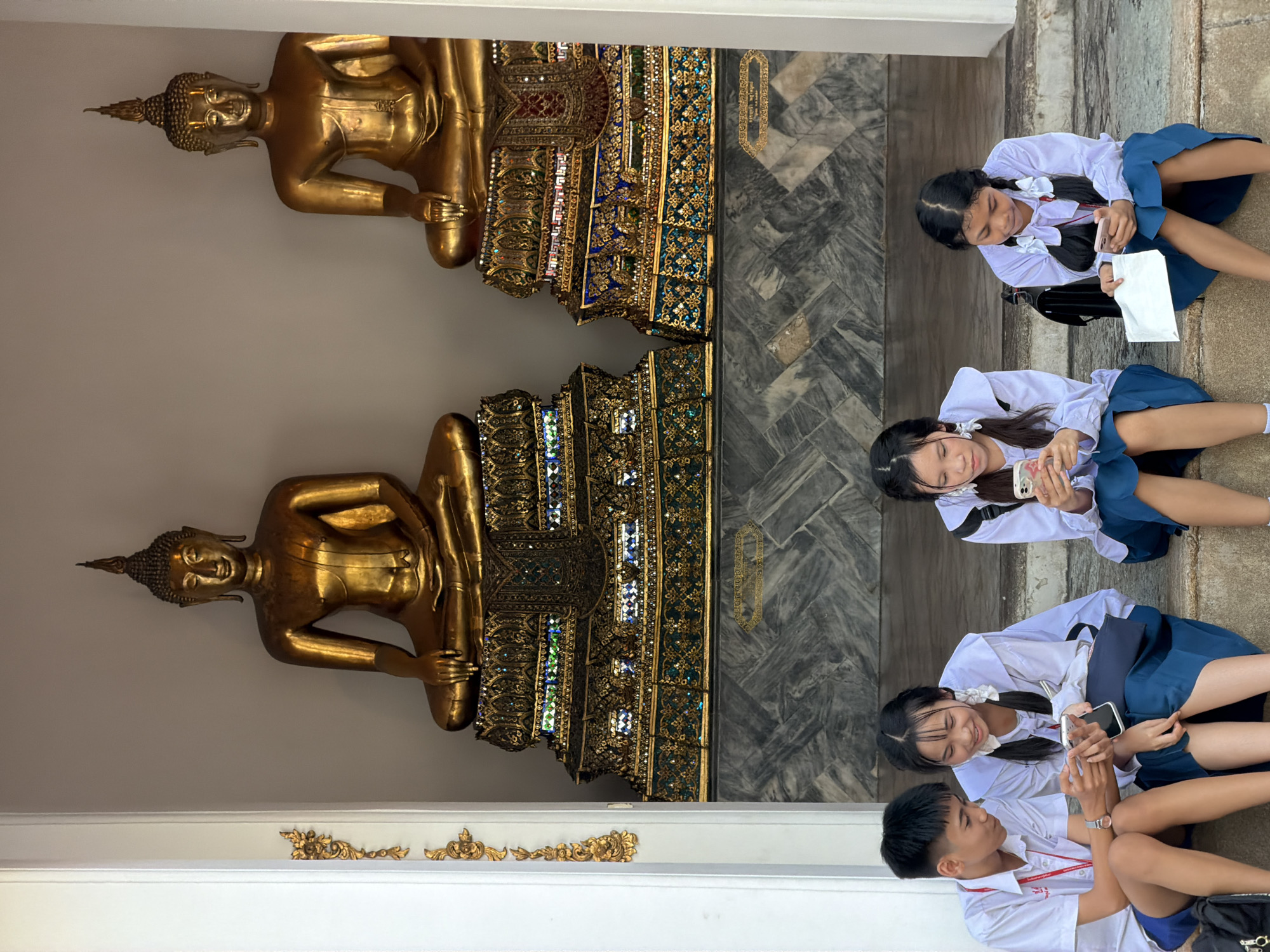 Four students, two males and two females, are seated on steps in front of two large golden Buddha statues. The students are wearing school uniforms consisting of white shirts and blue skirts or shorts. All four are looking at and interacting with their mobile phones. The Buddha statues are seated in meditation poses on ornate, jewel-encrusted pedestals. The background appears to be an open-air structure with marble-patterned walls. This scene is likely at Wat Pho (Temple of the Reclining Buddha) in Bangkok, Thailand, a well-known tourist attraction.