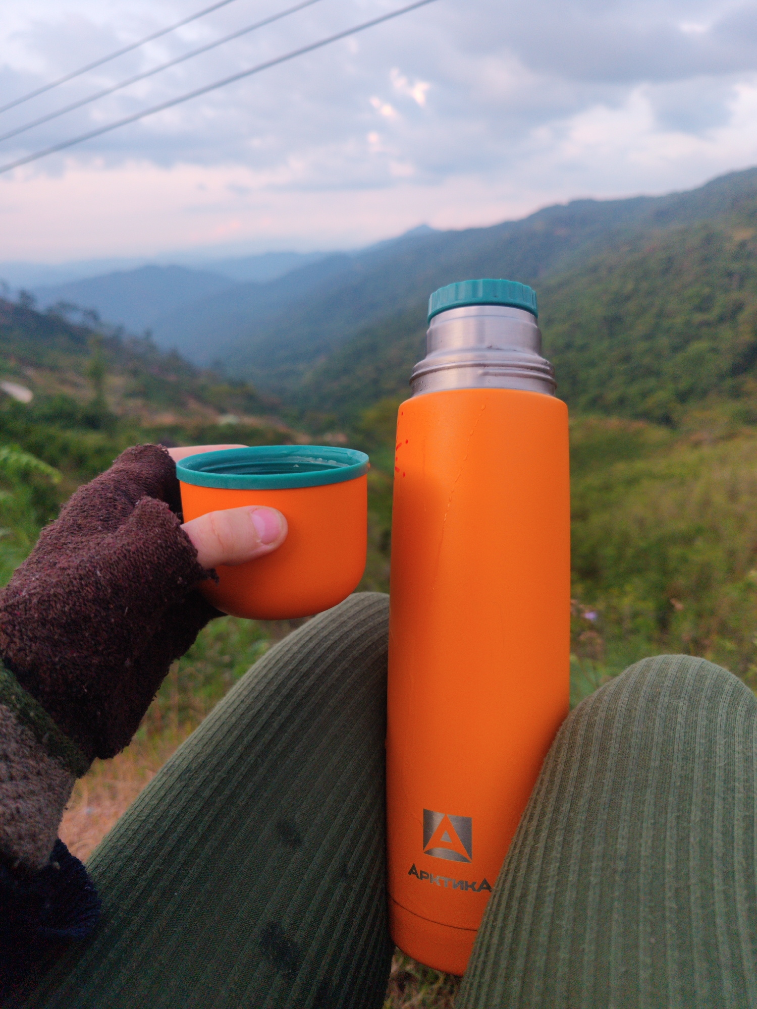 A person sitting outdoors in a mountainous region is holding an orange cup filled with liquid in their gloved hand, next to an orange thermos with a green lid. The person is wearing green corduroy pants and a brown glove. The thermos has a silver logo on its base. In the background are green hills and mountains under a cloudy sky. The location is indicated as Dam Rong 4, Vietnam. Power lines are visible above the mountainscape.