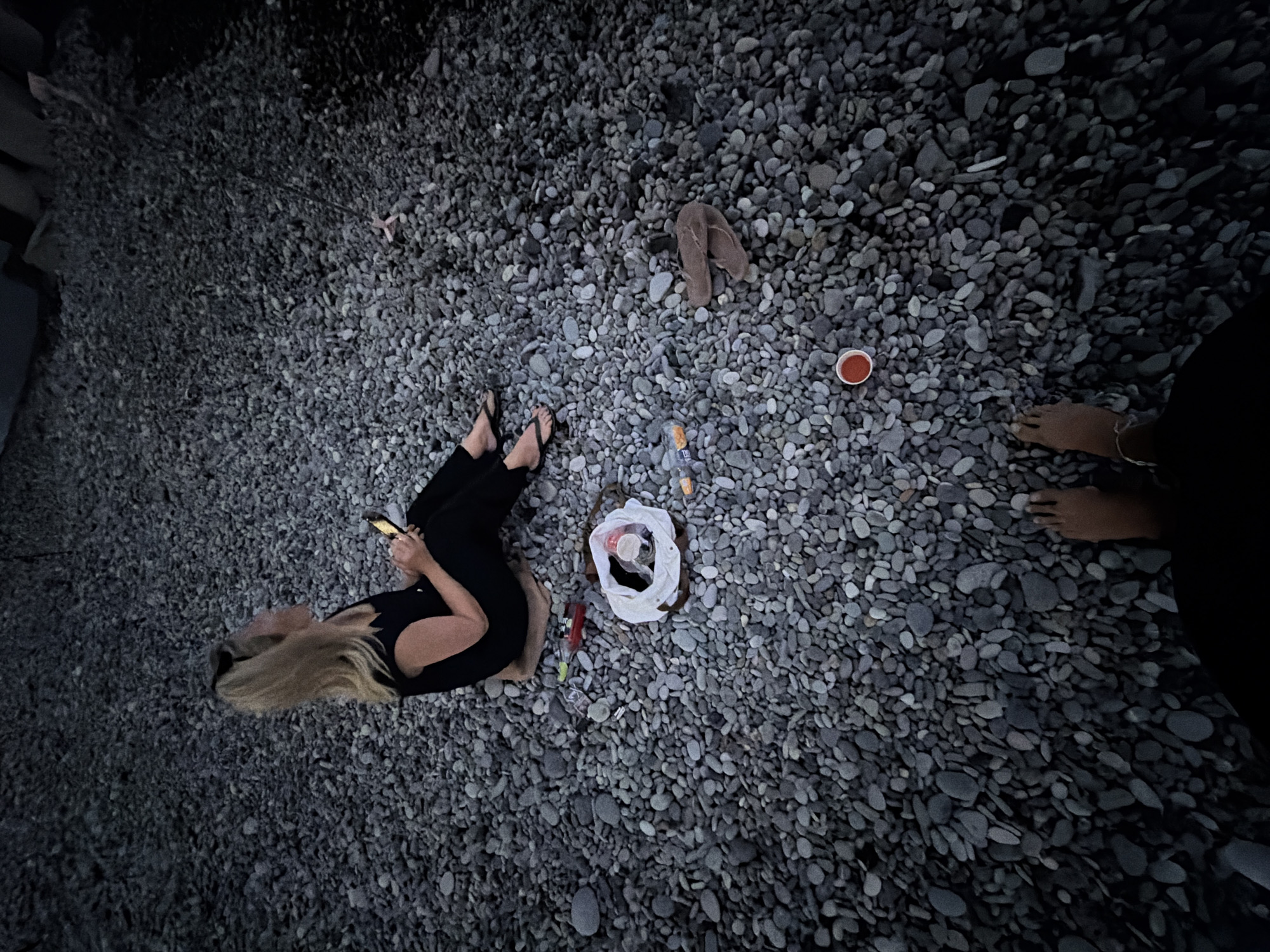 The image shows a person sitting on a pebble beach with a smartphone in hand. The setting appears to be dusk or night, with low lighting conditions. There are various items scattered on the pebbles, including a plastic bag, a red cup, and a pair of flip-flops. The person is wearing a black top and pants, and appears to be looking away from the camera. The background suggests an outdoor environment, possibly a coastal area. Given the location of Sochi, Russia, this scene could be at one of its many pebble beaches along the Black Sea coast.