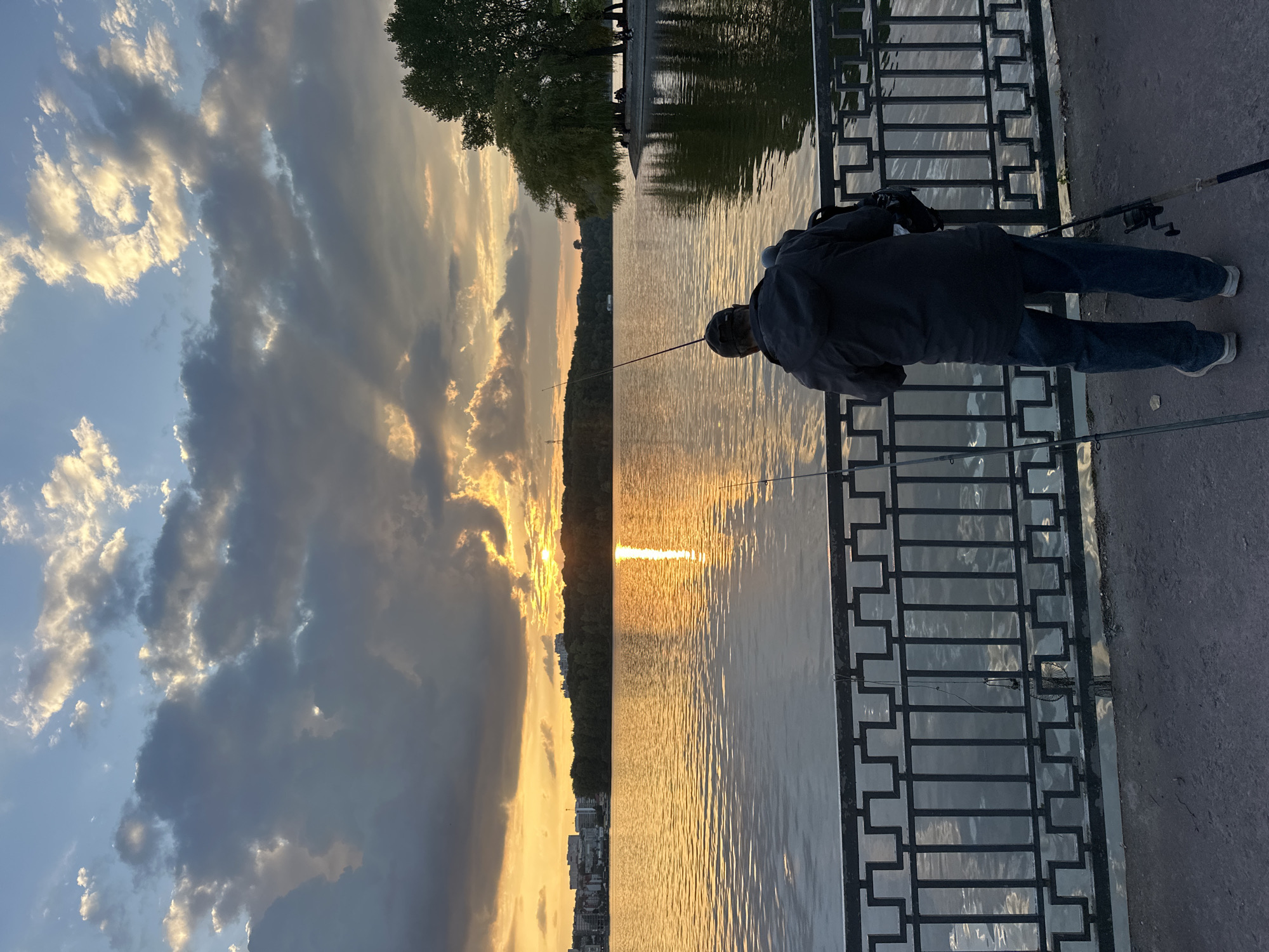 An individual stands on a paved walkway, facing a body of water, casting a fishing line. The person is wearing a dark jacket and jeans, with a baseball cap on their head. Two fishing rods are visible, one held by the person and another set up nearby. A metal railing separates the walkway from the water. The sun is setting, casting a warm golden glow across the water and illuminating dramatic clouds in the sky. In the distance, a treeline borders the far side of the water, with buildings visible beyond the trees. The setting is likely Ternopil, Ukraine, given the urban waterfront environment and the presence of a lake or large body of water.