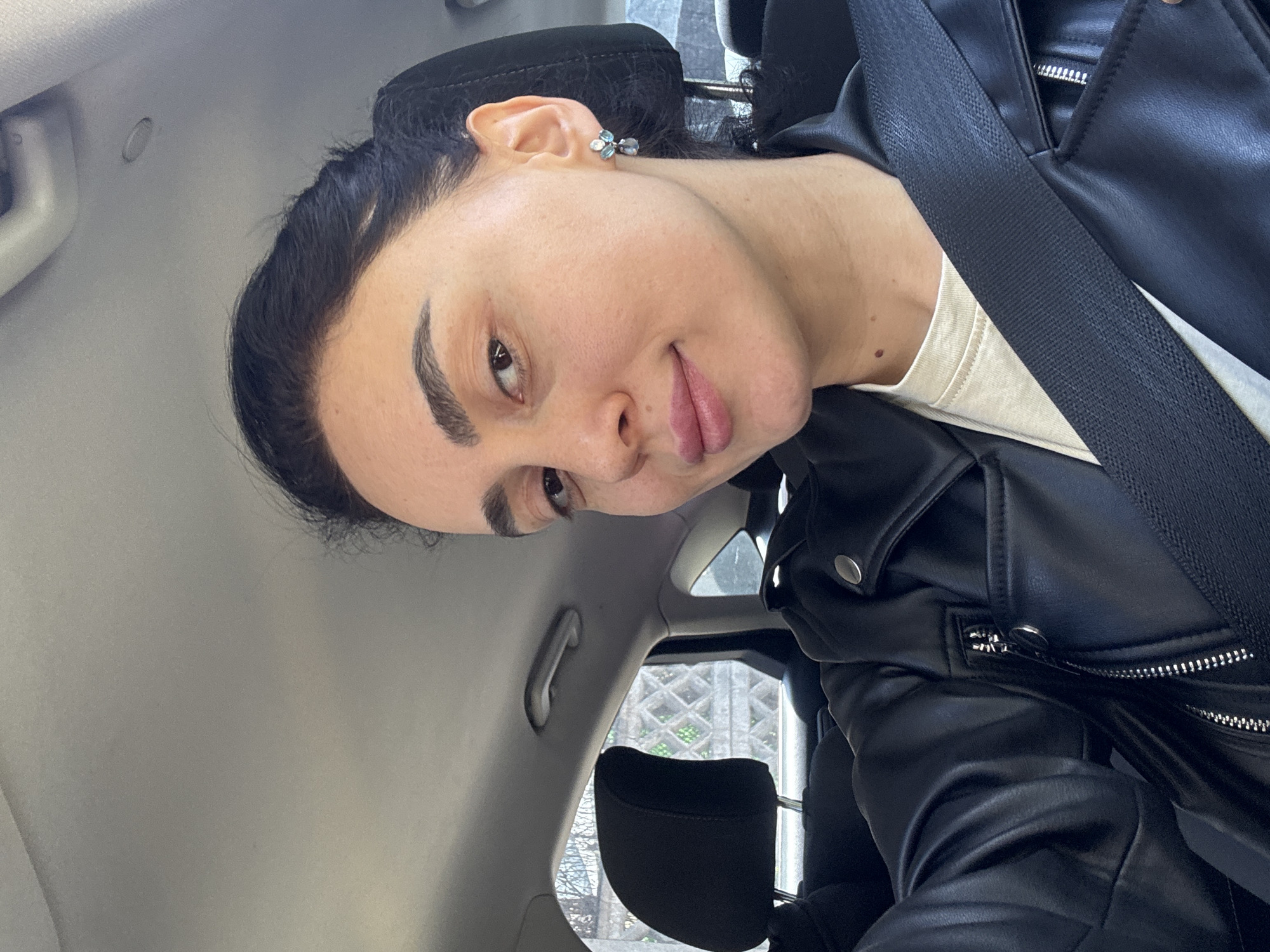 A woman is in a car wearing a black leather jacket and a white shirt. She is wearing a seatbelt and facing the camera. The location is Tbilisi, Georgia, as evidenced by the distinct architectural patterns visible through the car window. The interior of the car is predominantly gray.