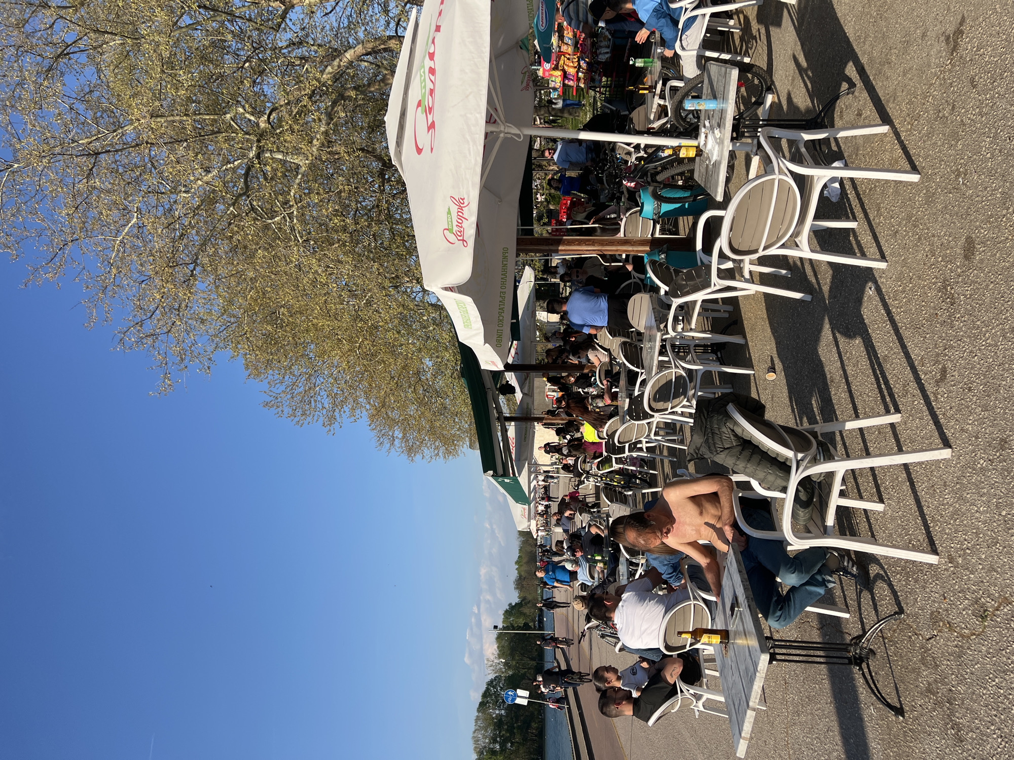 The image shows an outdoor scene in Plovdiv, Bulgaria. A long row of tables and chairs with white metal frames are set up, likely part of a café or restaurant. People are seated at the tables, some are wearing casual clothing. One man at a table is shirtless. Many bicycles are parked near the tables. A large white umbrella provides shade. The scene is alongside a waterfront walkway. In the background is a line of trees separating the walkway from the water. The sky is clear and blue. The weather appears to be sunny and warm.