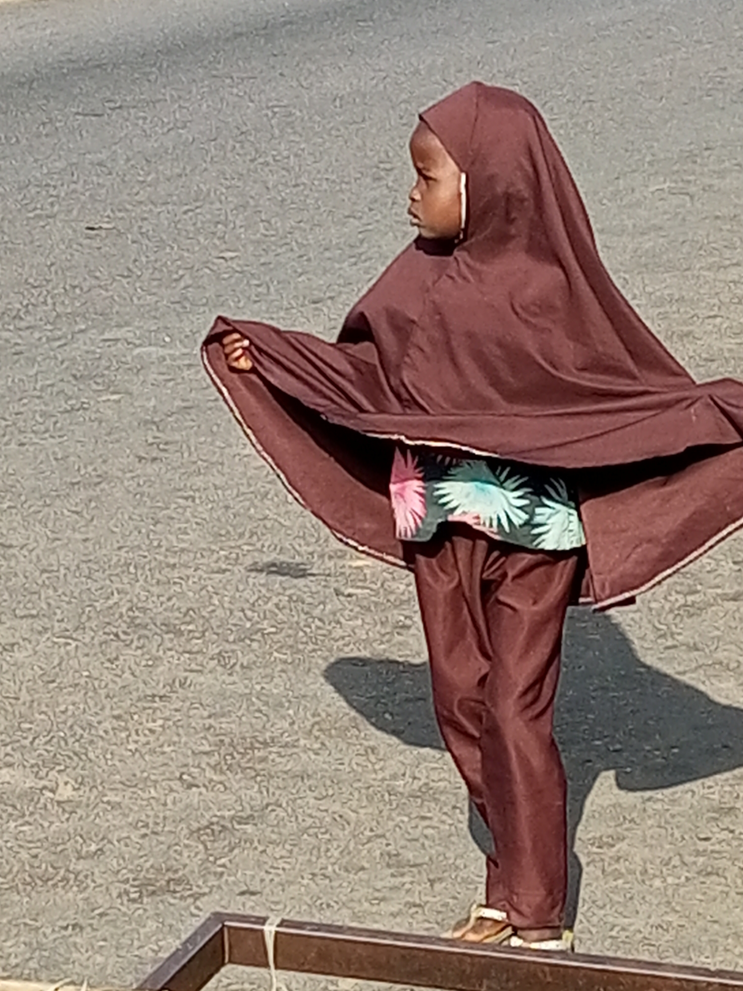 A young girl, appearing to be of African descent, is captured in profile, standing on a flat, rough-textured surface, possibly pavement or a dirt road. She is wearing a dark brown hijab that covers her head and extends down, creating a flowing cape-like effect. Beneath the hijab, a glimpse of a brightly colored shirt with a floral or abstract pattern in pink, green, and white is visible. She also wears dark brown trousers. Her right hand is raised, holding the edge of the hijab, as if adjusting it or caught in a gust of wind. The girl's gaze is directed to her left. The background is a uniform gray, suggesting an outdoor urban environment, potentially in the city of Tempure, Nigeria, given the context of the request. A portion of a brown metal structure, possibly part of a fence or barrier, is visible in the lower foreground, with white string tied to it. The lighting suggests it is daytime, casting a distinct shadow to the girl's right.