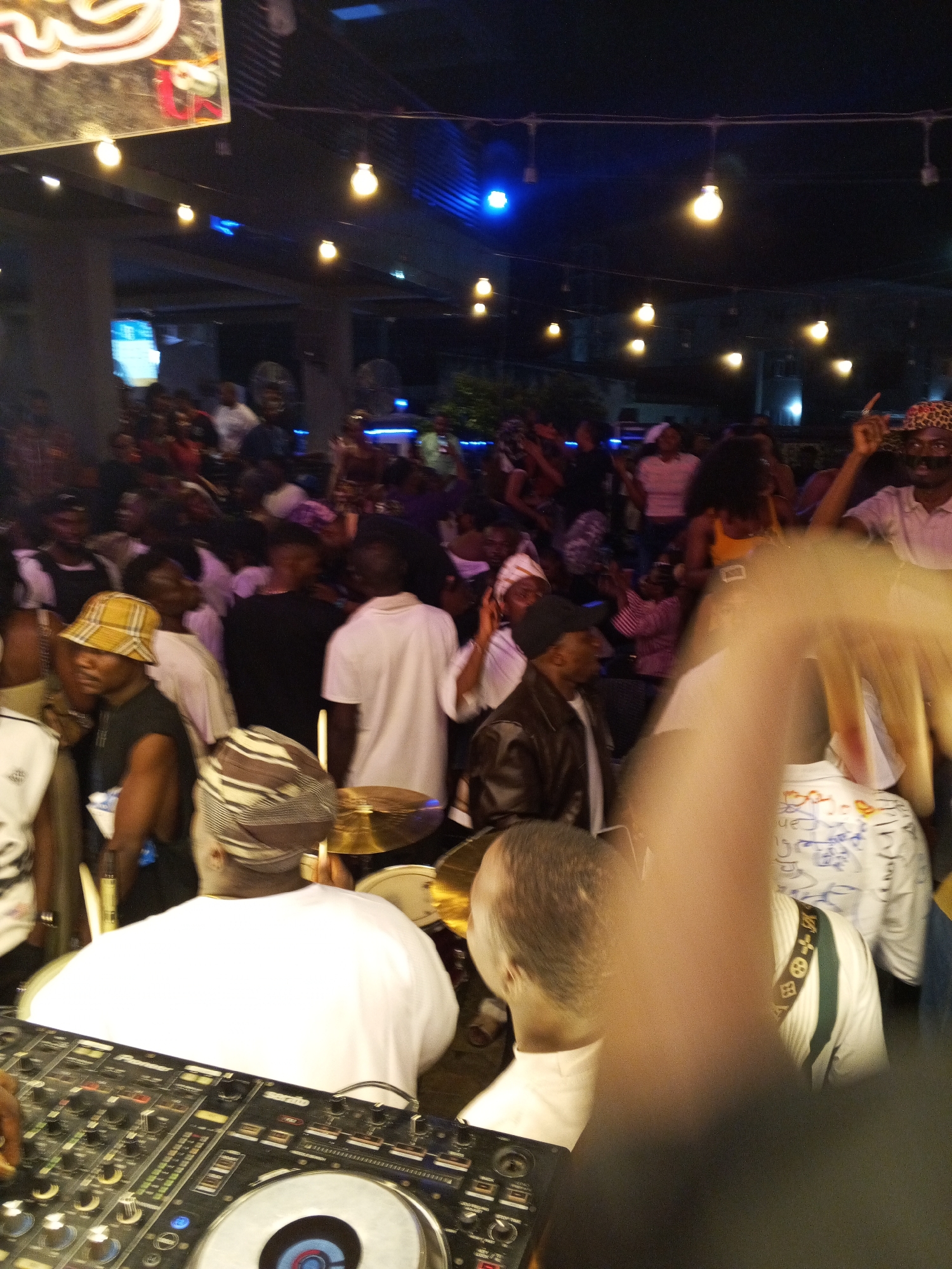 The image depicts a lively outdoor gathering at night, likely a party or event, in Lokoja, Nigeria. A DJ is visible in the foreground, operating a Pioneer DJ console. Behind the DJ, a person wearing a patterned head wrap is seen from the back, with drums and cymbals of a drum kit visible to their right. A crowd of people are gathered in the background, many of whom appear to be dancing or socializing. The scene is illuminated by string lights and spotlights, creating a festive atmosphere. The overall impression is one of a vibrant social event taking place in an urban setting.