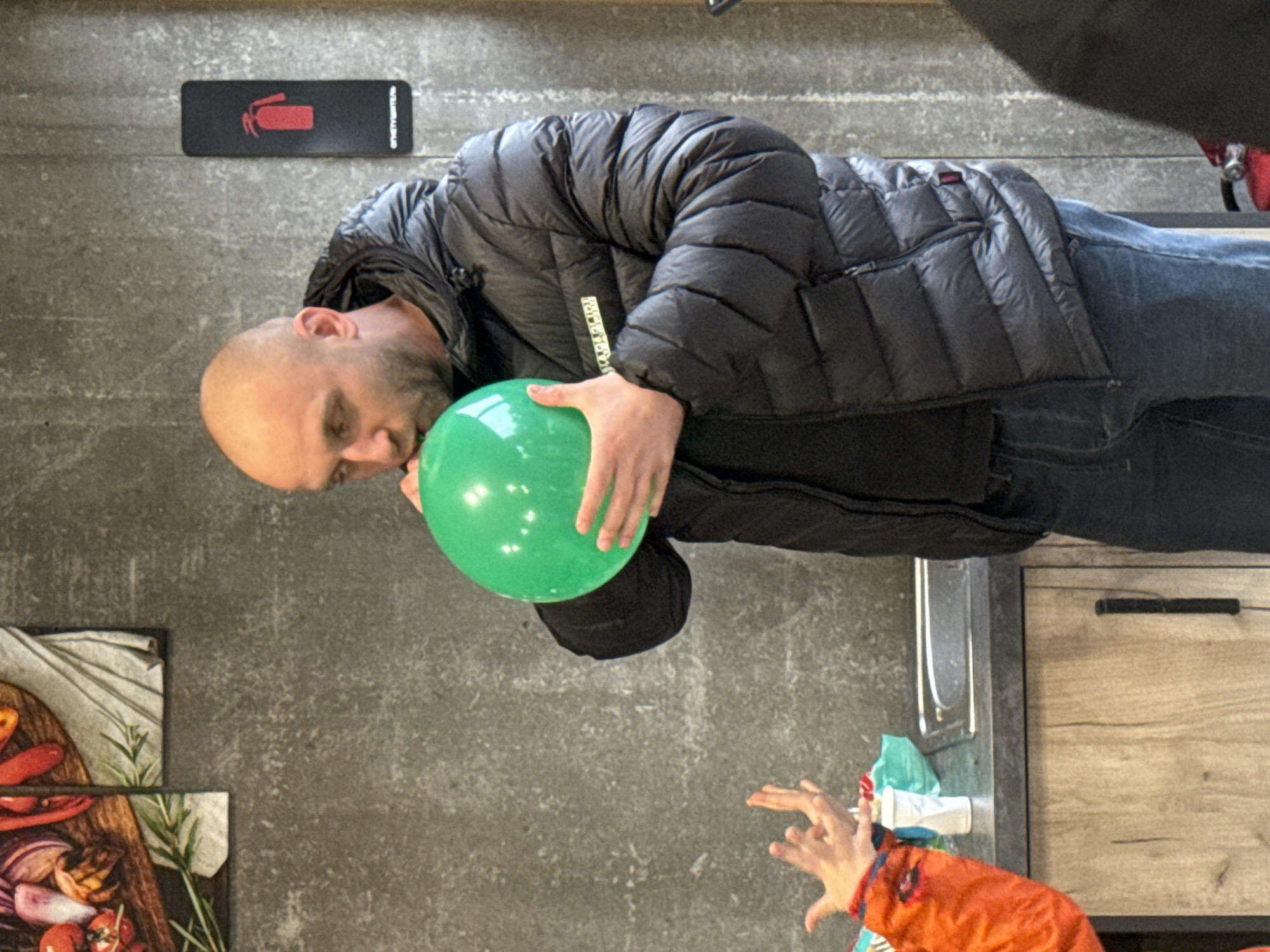 The image captures an indoor scene featuring a man interacting with a green balloon, while a child's hands reach out in the foreground. The image is oriented vertically, though the subjects appear to be standing or sitting upright in a conventionally viewed space.
The primary subject is a bald man with a beard, wearing a black puffer jacket, possibly a Napapijri brand jacket (indicated by visible text), and blue jeans. He holds a bright green balloon to his mouth, appearing to inflate it or playfully engage with it. His expression is one of focused attention.
In the lower right foreground, a pair of small hands, likely belonging to a child, are visible. The hands are outstretched, possibly reaching for the balloon, and the child wears an orange sleeve.
The background consists of a concrete or textured gray wall. Mounted on this wall is a black rectangular sign with a red fire extinguisher icon and the Russian word "ОГНЕТУШИТЕЛЬ" (FIRE EXTINGUISHER) in white text. To the lower left, part of a graphic or print depicting various vegetables such as red chilis, onions, and rosemary on a wooden cutting board is visible. To the lower right, a section of a light wooden cabinet or drawer with a black handle can be seen.
The setting appears to be a casual indoor environment, potentially a restaurant, cafe, or common area, consistent with the fire safety sign and interior decor elements. The scene suggests a lighthearted, playful moment between the man and the child. The location context is Yekaterinburg, Russia, which aligns with the Russian text on the fire extinguisher sign.