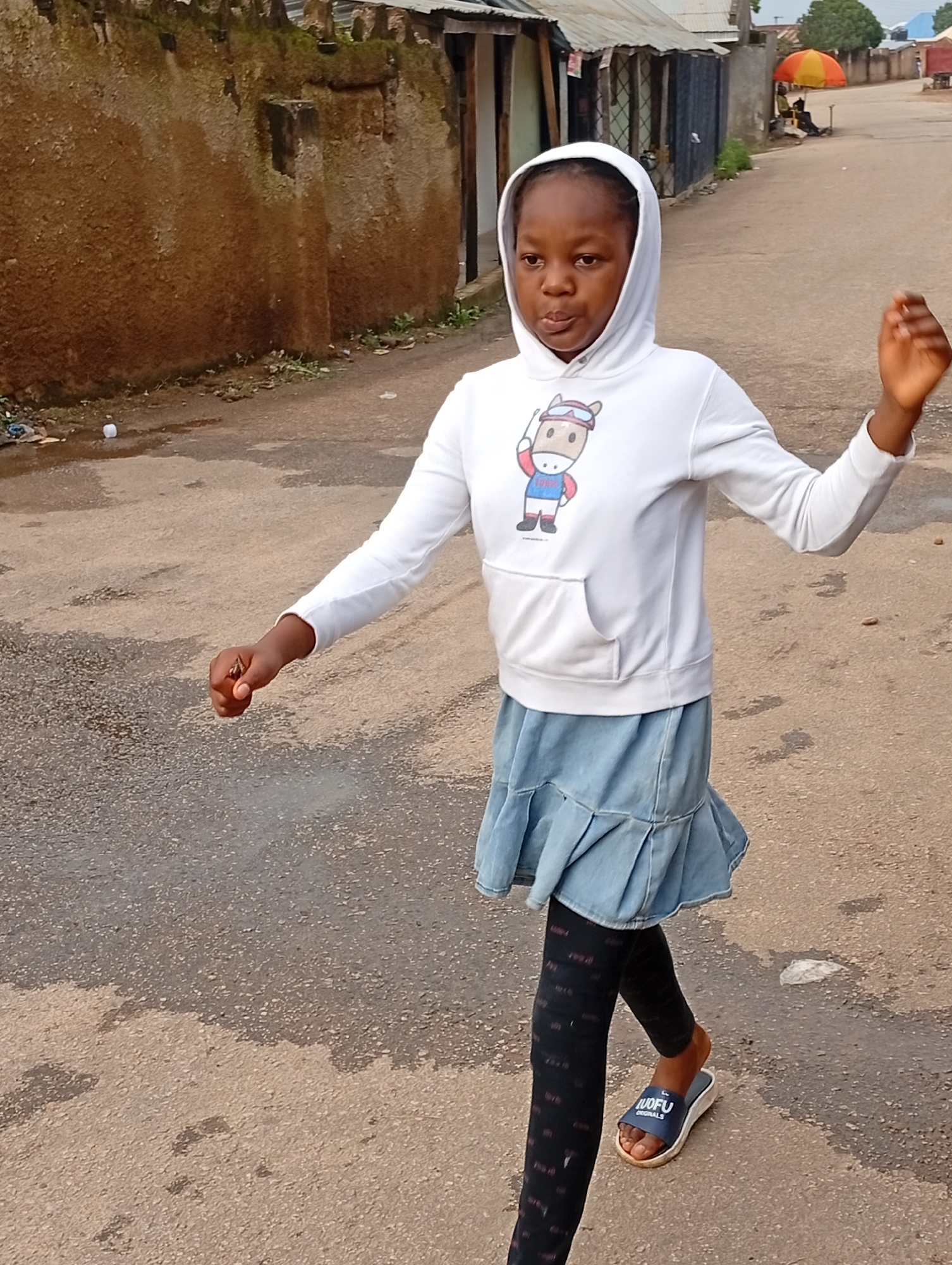 A young girl, likely pre-teen, with dark skin and braided hair, is captured in a medium shot outdoors. She is wearing a white hooded sweatshirt with a cartoon image of a horse on the front, a light blue denim skirt with a ruffled hem, black leggings with a repeating red pattern, and blue slides. The girl is in motion, with her right arm extended and her left arm bent at the elbow, as if walking or running. She is on a paved road with wet patches, indicating recent rain. To the left of the girl, a rough, brown wall is visible, covered in patches of green moss. In the background, along the road, are several low-rise buildings with corrugated iron roofs and some structures that appear to be market stalls. An orange and yellow umbrella is set up near one of the stalls, with a person seated underneath. The overall impression is an everyday scene in a residential or commercial area of Jos, Nigeria.