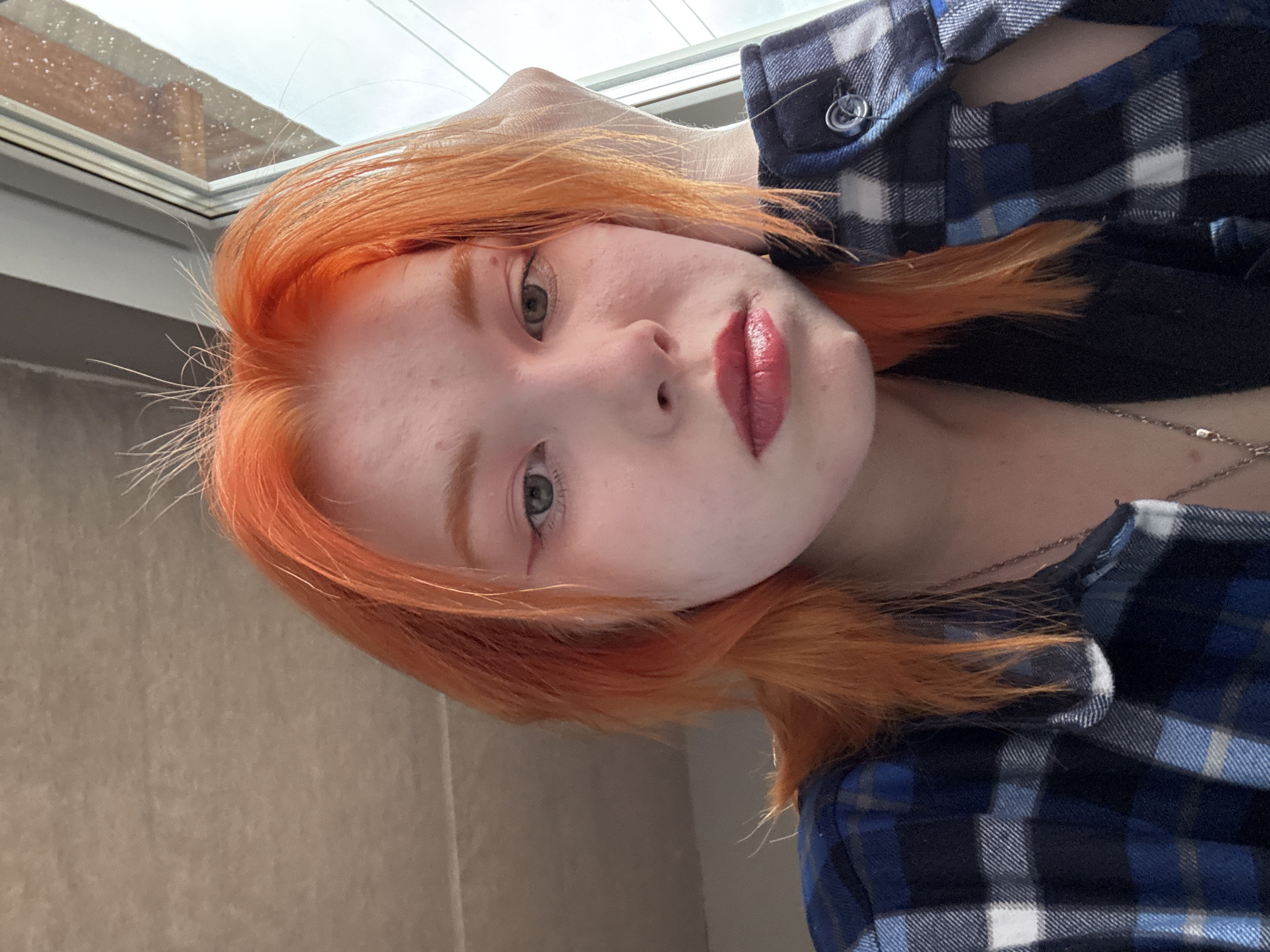 A close-up, slightly angled portrait of a young person with bright orange hair and pale skin, presumably female. She has light-colored eyes and wears red lipstick. A blue and white plaid shirt is visible. The background includes a window with visible raindrops and a beige wall. The location is Unknown, Unknown.