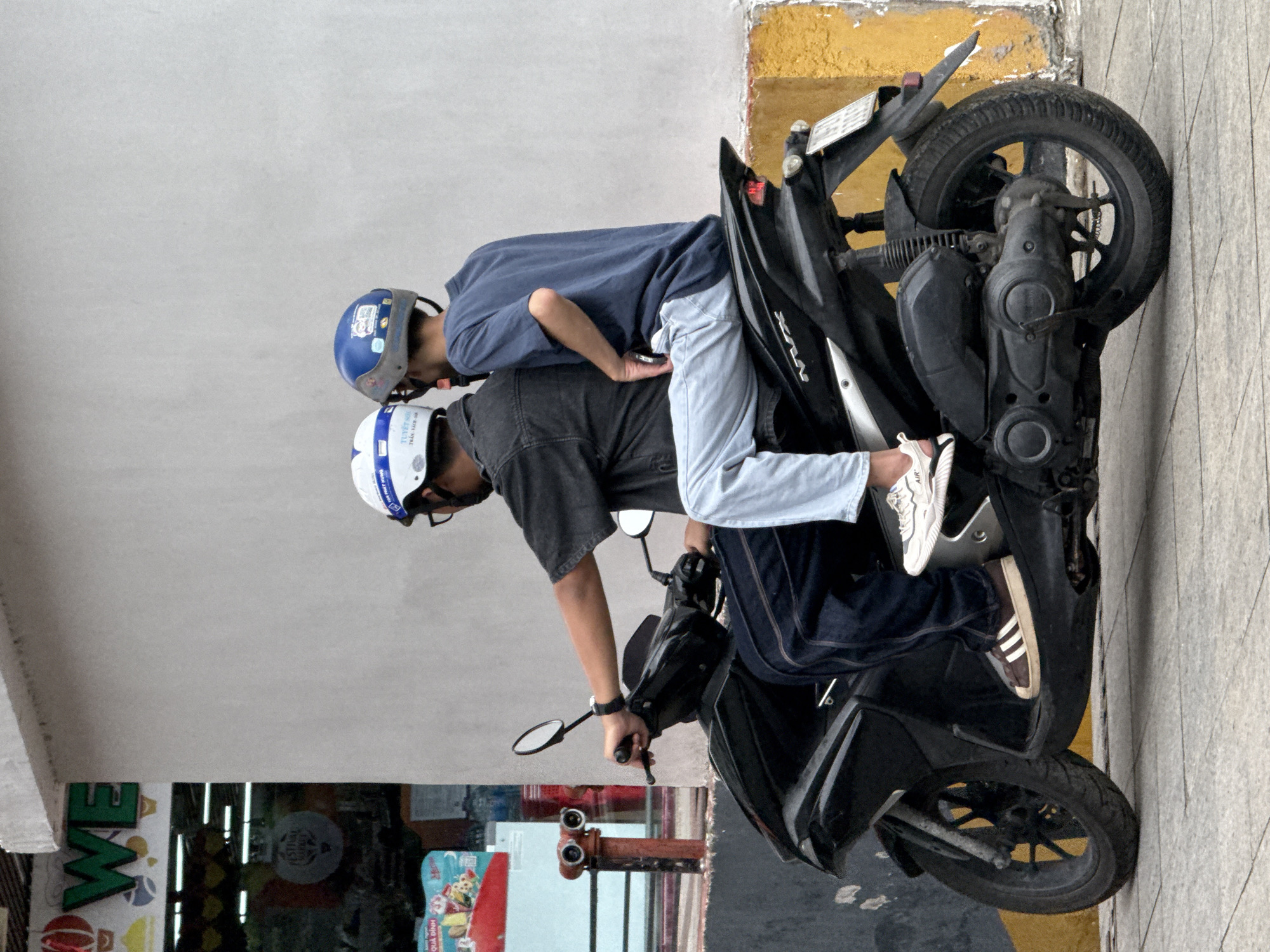 Two individuals wearing helmets are seated on a black motorcycle parked on a paved sidewalk. The person in front, wearing a white helmet with blue trim, is dressed in a dark denim jacket and dark blue jeans, with brown and white sneakers. The passenger behind, wearing a blue helmet with stickers, is in a navy blue t-shirt and light blue jeans. The motorcycle, a Yamaha Nvx model, is positioned in front of a building with a light gray concrete wall and a yellow and gray lower section. To the left of the motorcycle, a red fire hydrant is visible next to a storefront featuring a sign with green lettering that reads "WE" and a glass display window. The scene appears to be a street in Nha Trang, Vietnam, given the context provided.