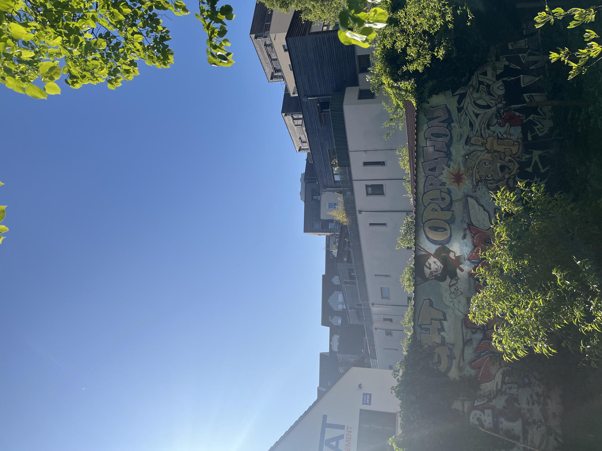 The media file presents a brightly lit outdoor urban scene under a clear, expansive blue sky. Vibrant green foliage from trees frames the upper left and right portions of the image, and also partially obscures elements within the scene.
On the left side of the frame, a series of modern, multi-story buildings with dark, possibly wooden or composite, facades and lighter-colored sections are visible, featuring balconies and multiple windows. To the right, a large wall is prominently displayed, covered with elaborate and colorful graffiti art. The graffiti includes cartoon-like characters, stylized letters, and an explosion or starburst motif. Legible text within the graffiti includes "OPERATION" and "CHT". This graffiti-covered wall is partially veiled by lush green foliage. Further down and to the right, a light-colored building is visible, displaying large letters that read "AT" and a partial "EMENT", likely part of a company or building name. A smaller blue sign on this building indicates "DEPUIS 1940".
The scene is bathed in direct sunlight, suggesting a clear and sunny daytime. A faint white speck, possibly a distant bird or airplane, is visible against the blue sky in the lower-left area. The overall setting combines elements of contemporary architecture, nature, and street art.