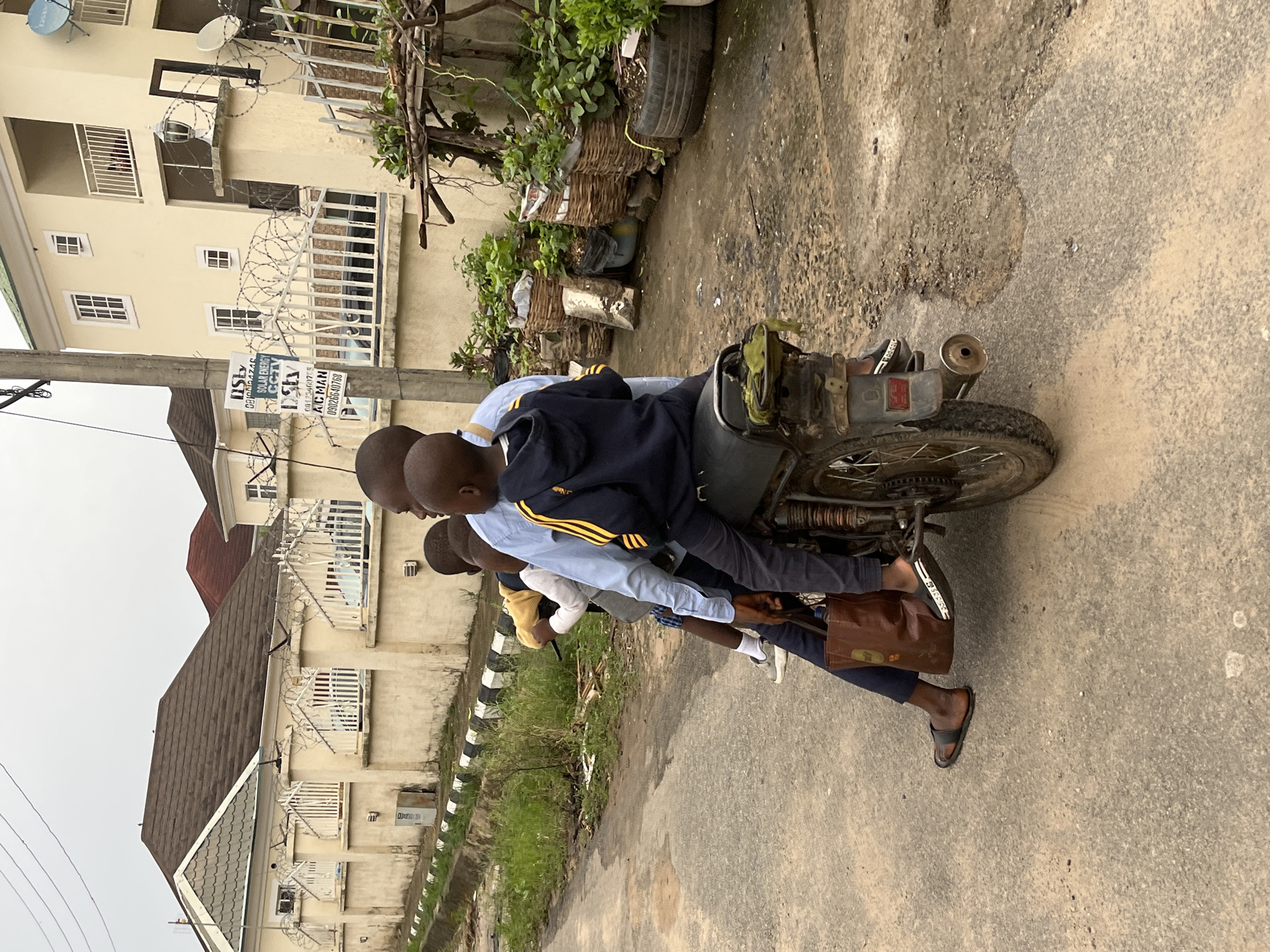 Three individuals are on a motorcycle on a street. The street is paved and has residential buildings on either side. The buildings are multi-story and some are surrounded by fencing with barbed wire. The individuals are dressed in casual clothing and appear to be riding together. The location is likely in a residential area of Abuja, Nigeria, based on the architectural style and street scene.