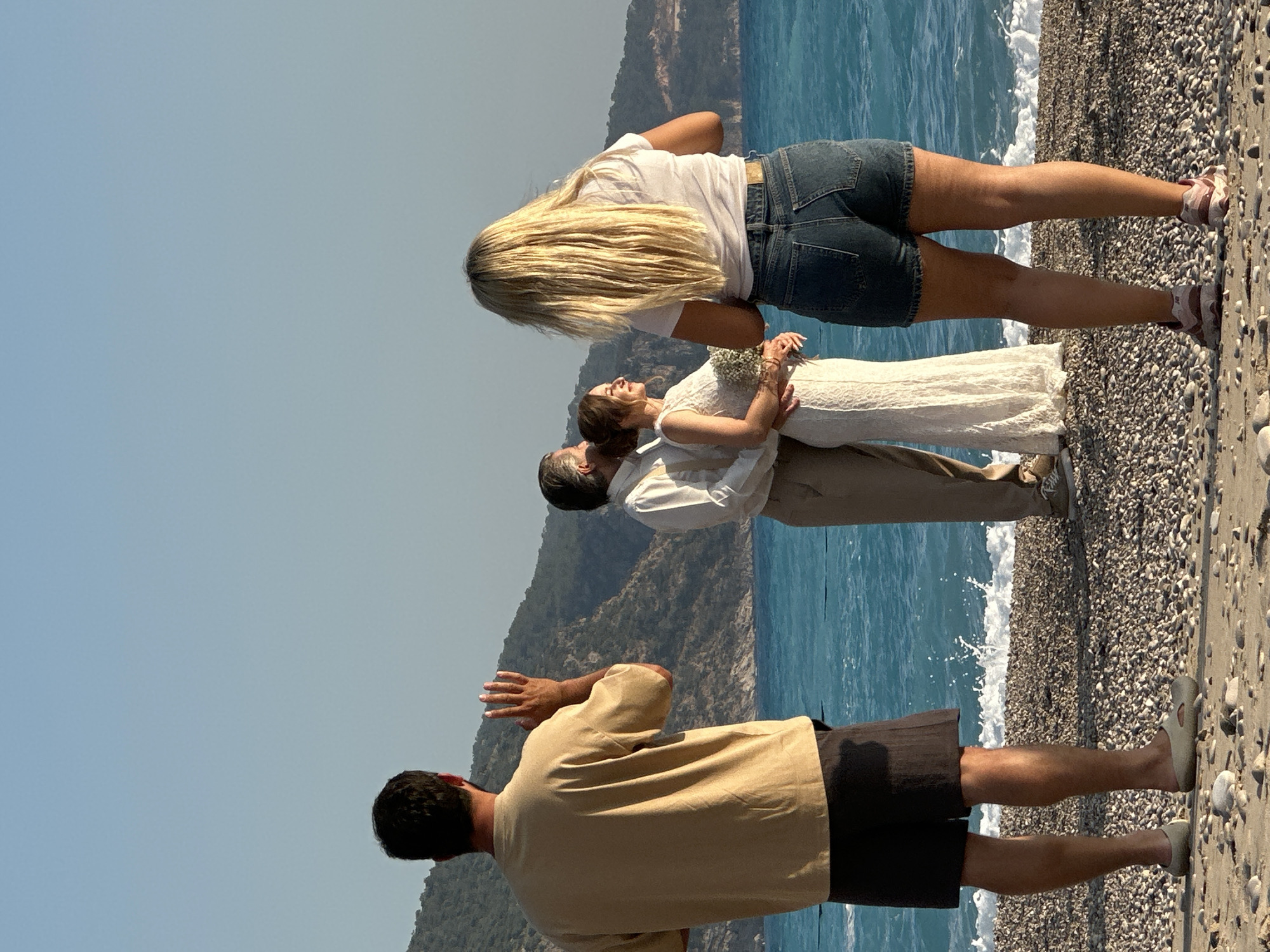 A couple is embraced on a pebble beach in Fethiye, Turkey, while being photographed by a man in a tan shirt and dark shorts. A woman with long blonde hair, wearing a white shirt and denim shorts, looks on. The beach is adjacent to turquoise water with small waves. Mountains are visible in the background, under a clear blue sky.