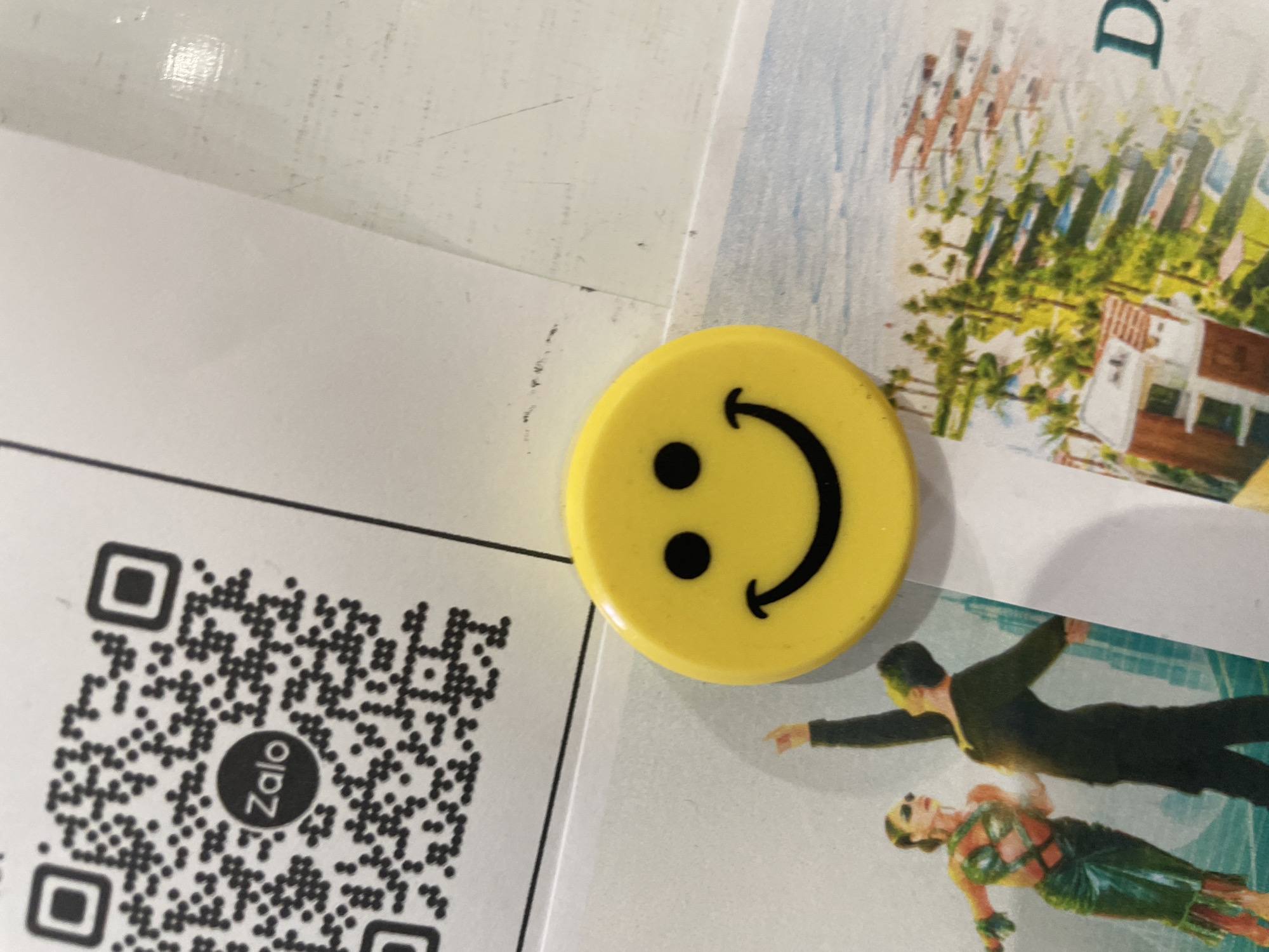 The image shows a close-up of a refrigerator door. A yellow smiley face magnet is attached to the door, obscuring part of a QR code. The QR code has the Zalo logo in the center. To the left of the magnet, a portion of a postcard or advertisement is visible, featuring two dancers. To the right of the magnet, another postcard or advertisement depicts a resort with palm trees and buildings. The background appears to be a white, possibly tiled, surface. The image is cropped to focus on these elements, and the context of Thủ Đức, Vietnam, is not visually present in the provided scene.