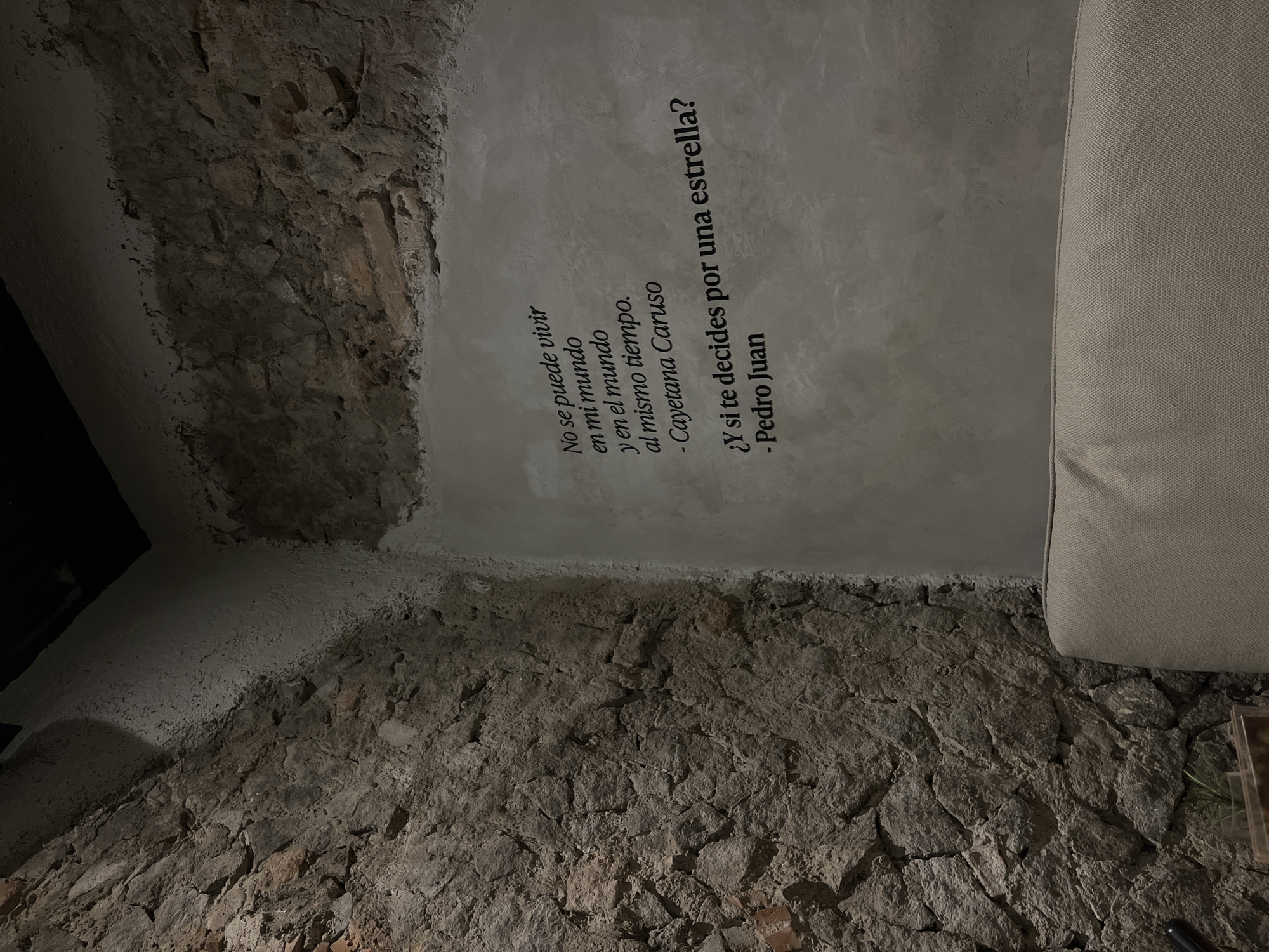 The image shows a section of a wall, likely indoors, featuring a combination of exposed stone and a plastered or cement-like surface. The lower portion of the wall is constructed of rough, uneven stones, giving it a rustic appearance. Above, a smoother, light-grey surface displays black text in Spanish. The text appears to be a quote or excerpt, with attributed names. A portion of a beige object (possibly a sofa or large cushion) is visible on the right. The overall lighting is dim, creating shadows and emphasizing the textures of the stone and the wall. The exact location in San Bernardino Tlaxcalancingo, Mexico is not discernible from the image.