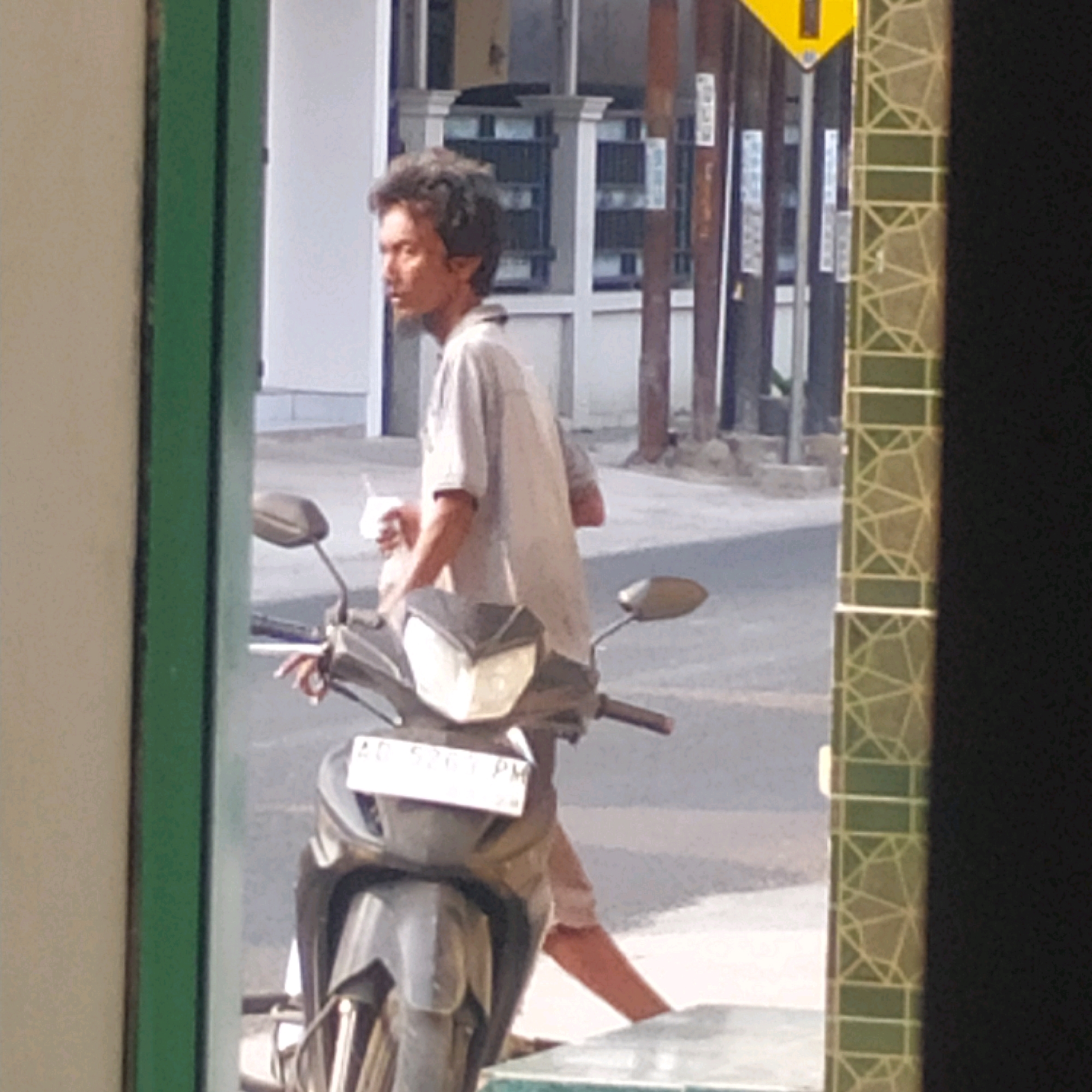 A young man with unkempt dark hair and a light-colored shirt is sitting on a dark-colored scooter in a semi-urban environment. He is holding a white plastic bag in his left hand and looking to his left. The scooter has a license plate that reads "AD 5261 PM". The background consists of buildings and possibly a street. The man's clothing and the overall scene suggest a casual setting.