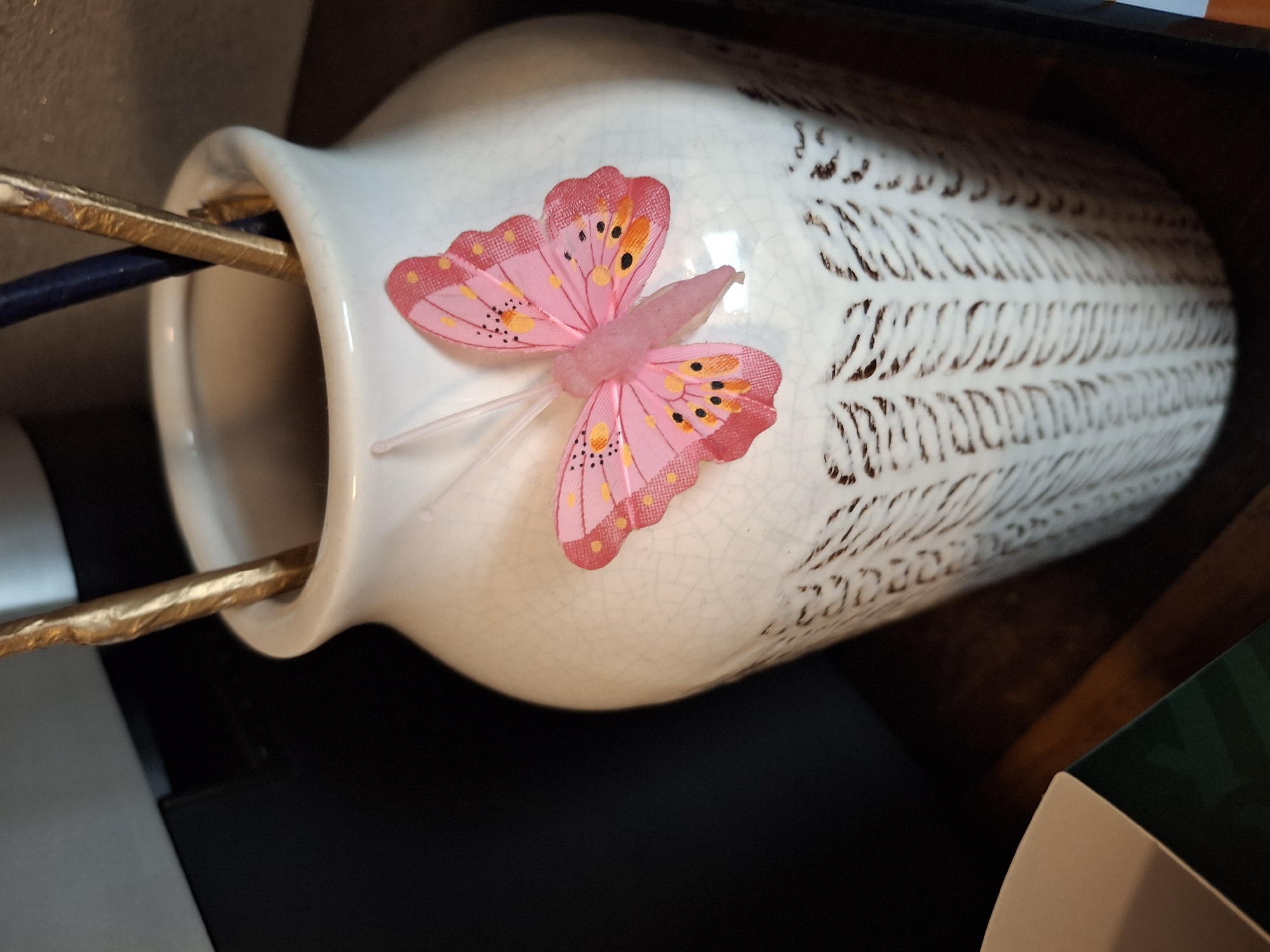 An ornate white ceramic vase with a crackle glaze finish sits on a dark wooden surface. The vase is decorated with an intricate raised pattern in a darker color. A pink fabric butterfly with black and orange spots and clear antennae is attached to the front of the vase. Inside the vase, two thin, metallic gold-colored objects and a dark blue object, possibly decorative sticks or brushes, are upright. In the background, a dark, indistinct object, possibly an appliance, is visible to the left. The scene is lit from the front and right, casting shadows. The image appears to be a close-up shot of the vase and its contents. There is no information or visual cue in the image that indicates the location is in Lēdmane, Latvia.