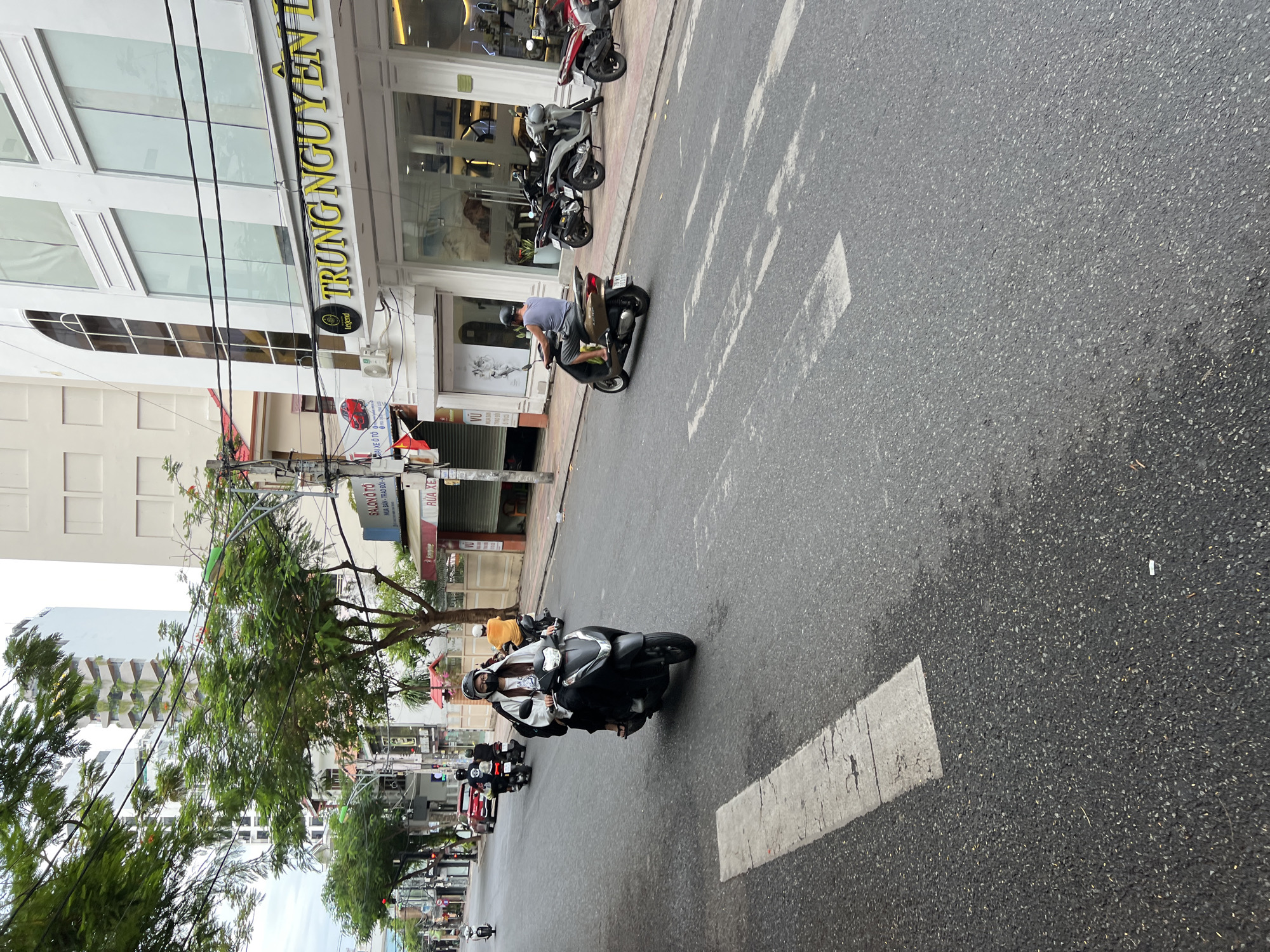 The image shows a street scene in Nha Trang, Vietnam, with several motorbikes on the road and a row of shops on the right. A young woman wearing a mask and a white jacket is riding a black scooter in the foreground, facing forward. She has a black helmet on. Behind her, a man in an orange shirt is riding a motorbike. Further down the street, more motorbikes are visible, some with riders and some parked in front of the shops. The shops have large glass windows displaying merchandise. The buildings are modern, with white facades and large windows. The road is paved with asphalt and has white lane markings. Trees line the street on the left side of the image. The sky is overcast. A sign on one of the shops reads "TRUNG NGUYEN LEGEND".
