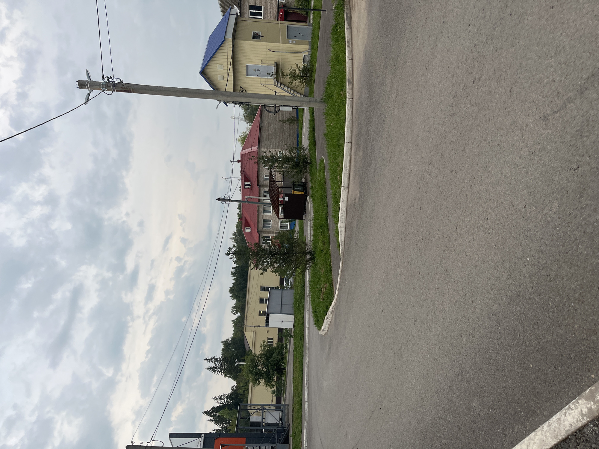 The image depicts a street scene in Blagoveshchensk, Russia, under an overcast sky. In the foreground, a grey asphalt road curves to the right, bordered by a white curb. A small circular patch of green grass with a white curb separates the road from the background.
Several buildings are visible in the mid-ground. To the left, a long, light yellow building with a flat roof and multiple windows is partially obscured by trees. Adjacent to it, a smaller grey utility structure stands. In the center, a building with a red tiled roof and visible windows is present. To the right, a narrow, two-story yellow building with a blue roof features an external staircase leading to an upper-level door. Another building with a red roof and windows is visible further to the right.
A concrete utility pole with wires and light fixtures stands prominently in the mid-right of the image. Trees, including evergreens, are scattered throughout the scene, particularly behind the buildings and along the roadside. A dark brown shelter with a dark colored bin and what appears to be a recycling bin are located near the red-roofed building. No people are visible in the image. The environment appears to be a semi-urban or industrial area.