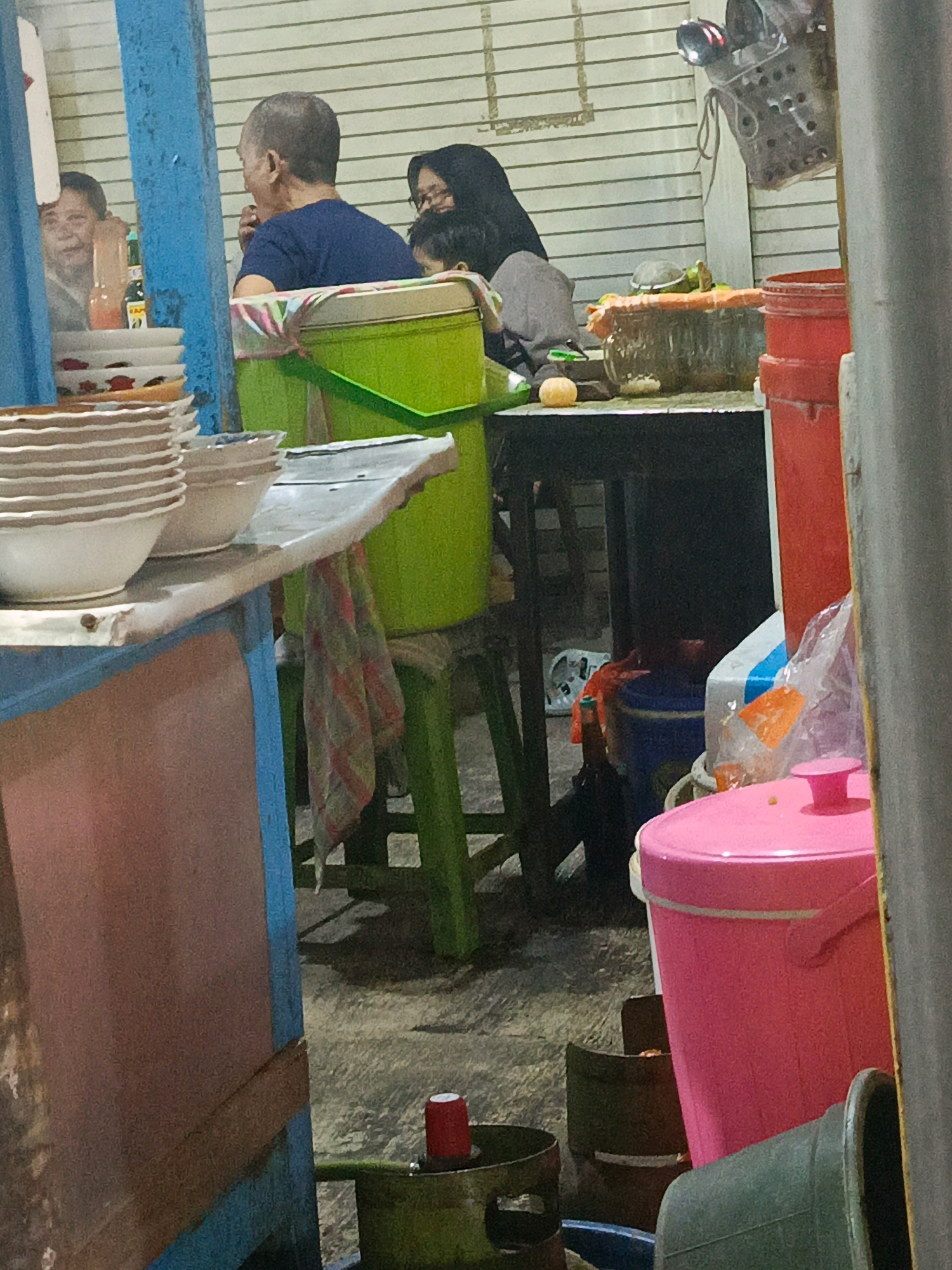 The image depicts an outdoor scene in what appears to be a food stall or market setting in Sedati Agung, Indonesia. In the foreground, a stack of white bowls with red floral patterns is visible on a blue and pink counter. To the right, a large bright pink plastic bucket with a lid is prominently displayed, accompanied by other containers and a green bucket with a red object on top. In the middle ground, a green plastic cooler is situated behind the counter, and a person wearing a dark blue t-shirt is seated at a table, facing away from the camera. Behind this person, another individual wearing a black hijab is visible, along with a young child. The background features a striped wall. The lighting suggests it might be evening or a shaded area.