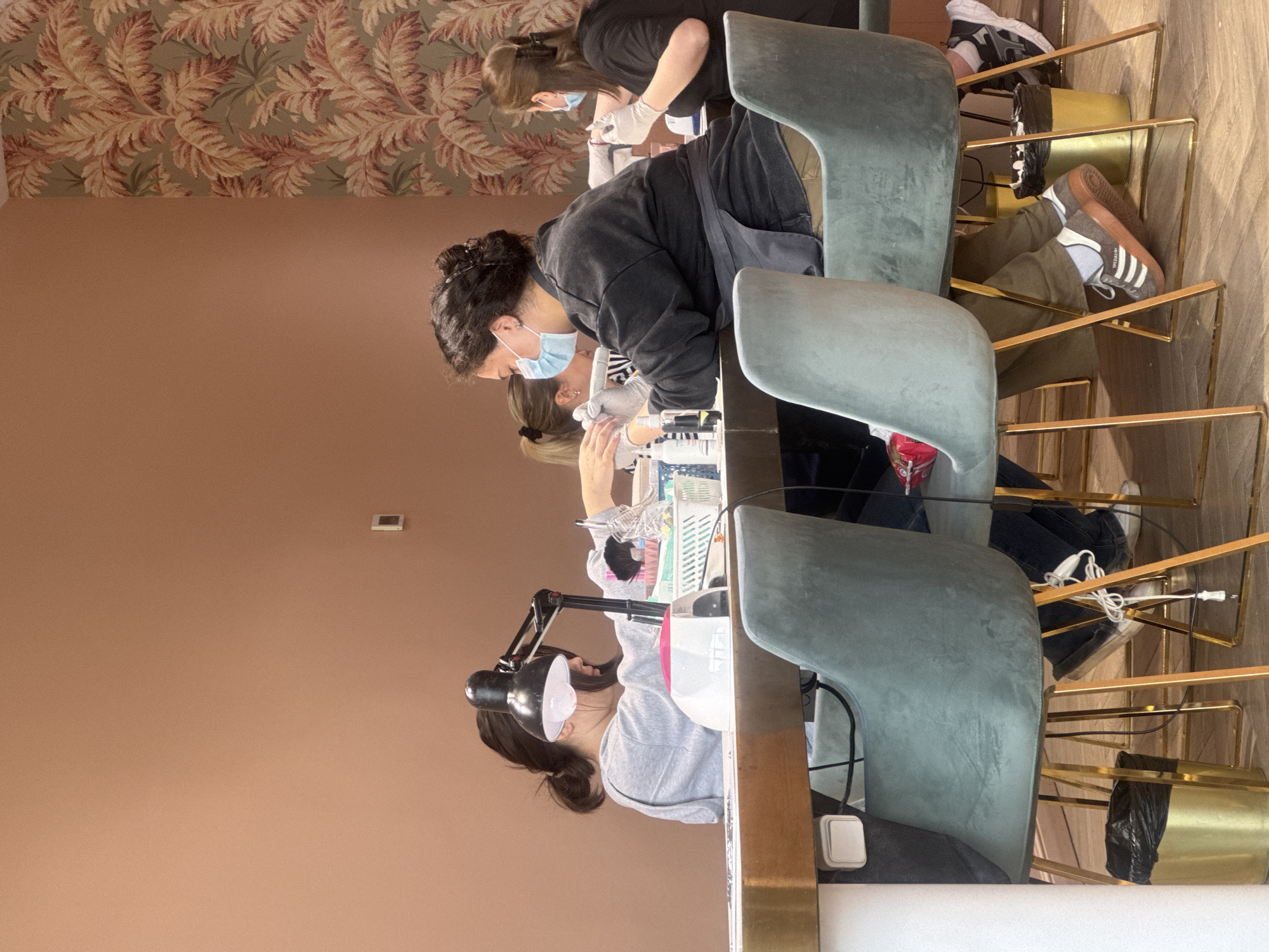 **Location:** The image appears to depict the interior of a nail salon. While the image does not contain location data, based on metadata analysis, it appears to be taken in Tbilisi, Georgia.
**Scene Description:** The image shows several individuals engaged in a nail care process.
* **People:** Multiple women are visible. Some appear to be nail technicians, while others seem to be receiving manicures. The technicians are wearing face masks and gloves, engaging in activities such as filing and painting nails. At least one woman is wearing Adidas brand sneakers.
* **Objects:** The scene features manicure tables, chairs, a lamp with a magnifying lens, and various nail care tools and products.
* **Actions:** The nail technicians are actively performing nail care procedures on the clients. The clients appear to be relaxing and receiving the treatments.
* **Environment:** The salon has a well-lit interior with a peach-colored wall and wallpapered ceiling. The decor is modern and designed to create a relaxing and hygienic atmosphere.
