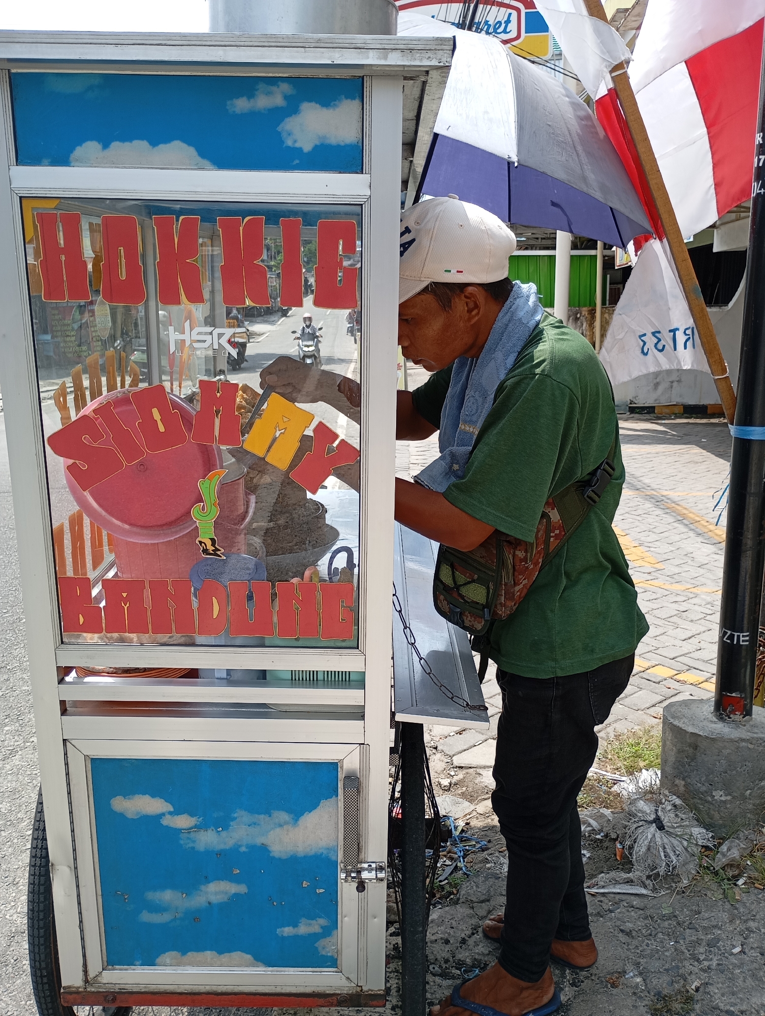 In Balikpapan, Indonesia, a man wearing a green t-shirt, black pants, and flip-flops stands beside a food cart. He is looking down into the cart, seemingly attending to its contents. The cart has a glass display with red lettering that reads "HOKKIC" and "SIOMAY BANDUNG" (a type of Indonesian steamed fish dumpling). The cart's lower section features a painted sky with clouds. In the background, a street with vehicles and a red and white Indonesian flag can be seen. The man has a light blue towel around his neck and a camouflage fanny pack slung across his chest. The cart is positioned on a paved surface, with some debris scattered around its base.