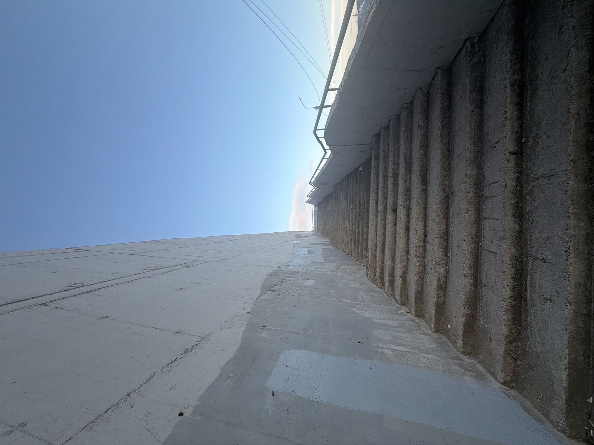 The image, taken in Krasnoyarsk, Russia, depicts a vast concrete structure viewed from a low, upward-looking angle against a bright, clear blue sky. On the left, a smooth, light grey concrete wall extends upwards, displaying horizontal and vertical seams, along with some patches of lighter grey, possibly from repairs or fresh paint. To the right, a darker, heavily textured concrete wall with prominent vertical ribs rises sharply, giving an impression of massive scale and robustness, typical of a dam or large retaining wall.
Above the junction of these two concrete surfaces, a metal railing is visible, topped by thin power lines stretching across the sky. A street lamp fixture can be seen attached to the upper edge of the structure. A strong light source, likely the sun, is visible high in the sky, casting a bright glow and suggesting sunny daytime conditions. No people or specific activities are present in the frame. The scene highlights large-scale, utilitarian architecture, consistent with infrastructure found in locations like the Krasnoyarsk Dam. There is no visible text.