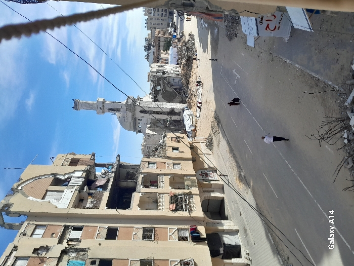 The image captures a street scene in Khan Yunis, Palestinian Territory, showing significant urban destruction under a clear blue sky. A multi-story building on the left exhibits extensive damage, with large sections of its facade missing, revealing exposed interiors, and laundry items hanging from balconies. In the mid-ground, a severely damaged mosque features a partially collapsed minaret and dome. The asphalt street below is marked with a white center line and a white directional arrow. A man in a white shirt and dark trousers walks leftward on the street in the foreground. Further up the street, two figures in dark clothing, appearing to be a woman and a child, walk away from the viewer. Multiple individuals are also discernible among piles of rubble in the background near the mosque. The right side of the street and background areas are littered with debris and show further damaged buildings. Overhead wires traverse the frame. A partially visible white sign with red Arabic script is present on the right foreground.