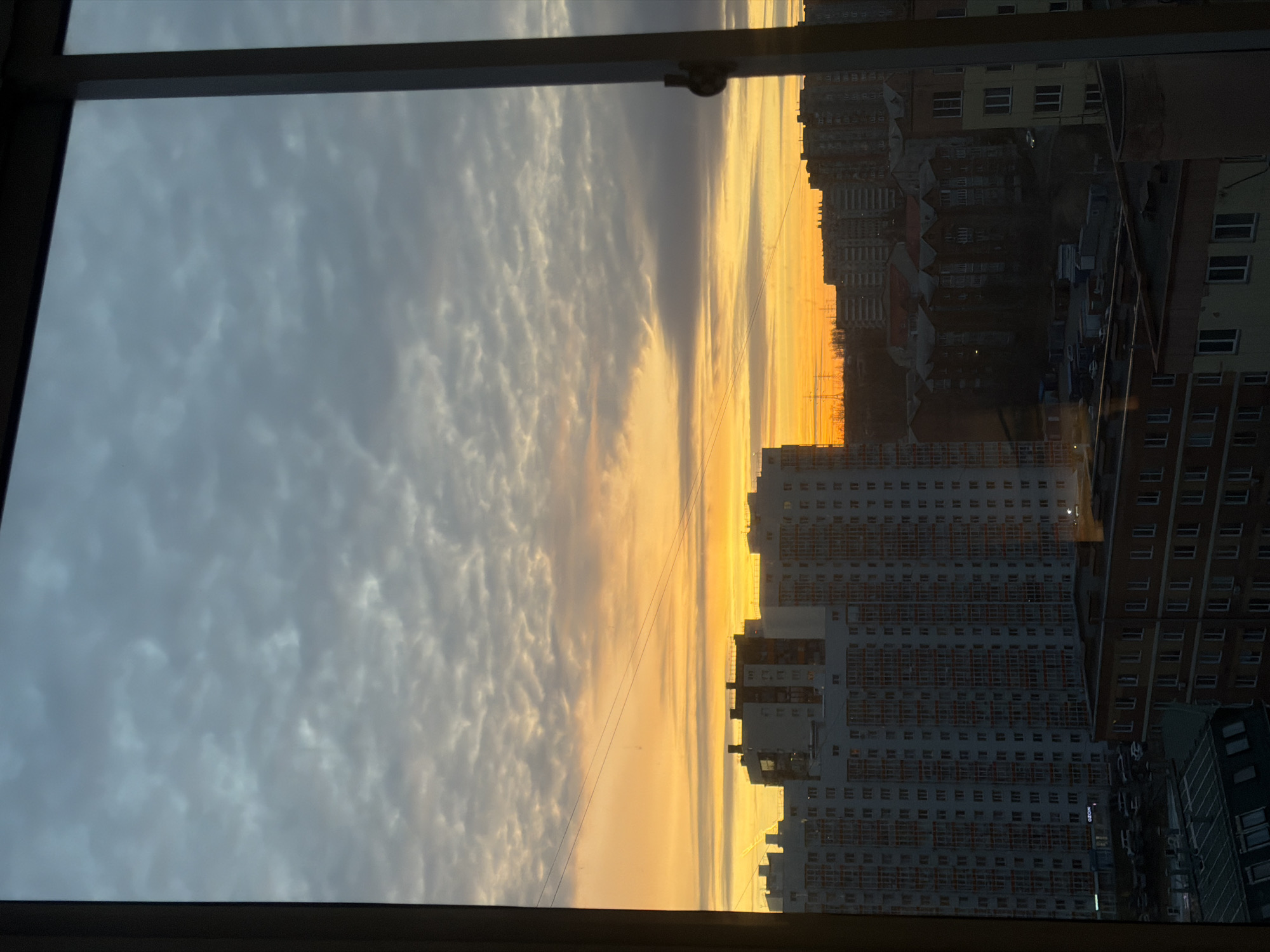 A low-angle indoor shot through a window captures a cityscape at sunset. The sky transitions from a cloudy gray on the left to a vivid orange near the horizon. Power lines stretch across the view. Several high-rise buildings are visible, their facades reflecting the setting sun. The buildings are primarily residential, with many windows. To the right, taller, denser buildings suggest a city center. The window frame is dark, creating a natural border around the scene. Based on the building style and the "OZON" sign on one of the buildings, this image is very likely taken in Krasnoyarsk, Russia.