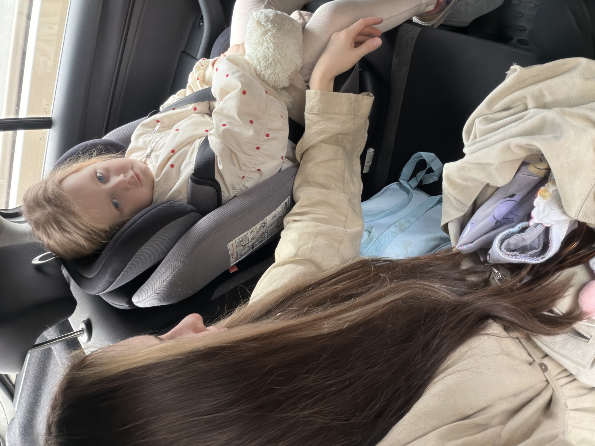 A young child is secured in a car seat within a vehicle, and an adult lies next to the child. The child has fair skin, blonde hair, and blue eyes, and wears a beige jacket with red polka dots. The car seat is gray and black, and the interior of the car is black. The adult has long, brown hair, and wears a beige jacket. Additional items in the car include a light blue bag and a pile of beige and purple clothing. The location is in a car, presumably in Khimki, Russia.