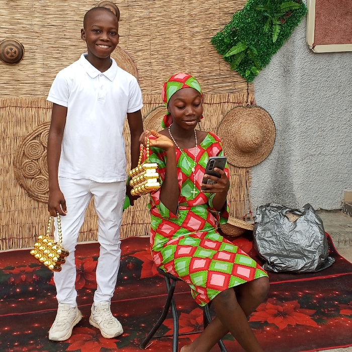 In Adankolo, Nigeria, two youths are depicted in a posed setting against a decorated wall and floor. A male youth stands on the left, facing forward with a slight smile. He wears a white short-sleeved polo shirt, white slim-fit pants, and white athletic shoes. In his left hand, he holds a small, rectangular, golden beaded bag with a string handle. A female youth is seated on a black folding stool to the right, her gaze directed downwards at a black smartphone held in both hands. She wears a colorful dress patterned with red, green, and light pink diamond shapes, complemented by a matching headwrap. A white rosary with a cross hangs around her neck. Her right hand holds an identical golden beaded bag, positioned near her lap. The background consists of a woven mat wall hanging featuring circular patterns, two straw hats, and a section of artificial green foliage. A grey textured wall is visible to the right. The floor is covered by a dark red rug adorned with scattered red floral patterns. A crumpled grey bag rests on the rug to the right of the seated female.