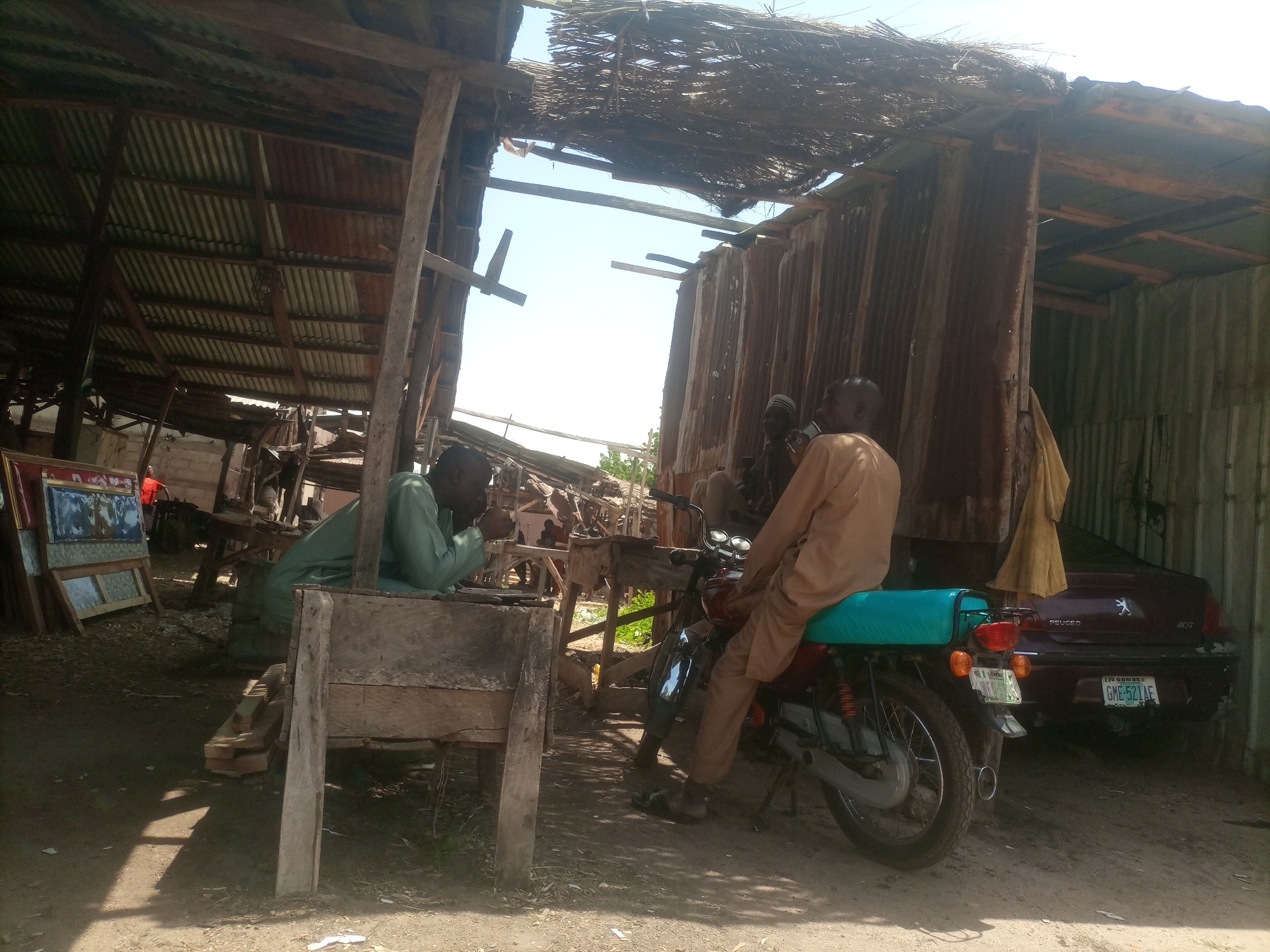 The image depicts a roadside scene under a makeshift shelter in Arawa, Nigeria. The shelter has a corrugated metal roof and walls, with some areas open to the sky. Two men are visible. One man, wearing a green garment, is seated behind a wooden workbench. He is looking down and appears to be engaged in some activity at the workbench. The other man, wearing a light brown robe, is seated on a motorcycle. The motorcycle is positioned in front of a dark red Peugeot 407 car, which is parked behind the motorcycle. The car has a Nigerian license plate that reads "GME-521AE". In the background, there are several framed pictures leaning against a wooden structure, and additional corrugated metal sheeting. The ground is dusty and uneven, with some scattered debris. The lighting suggests it is daytime, with sunlight filtering through the open parts of the shelter.