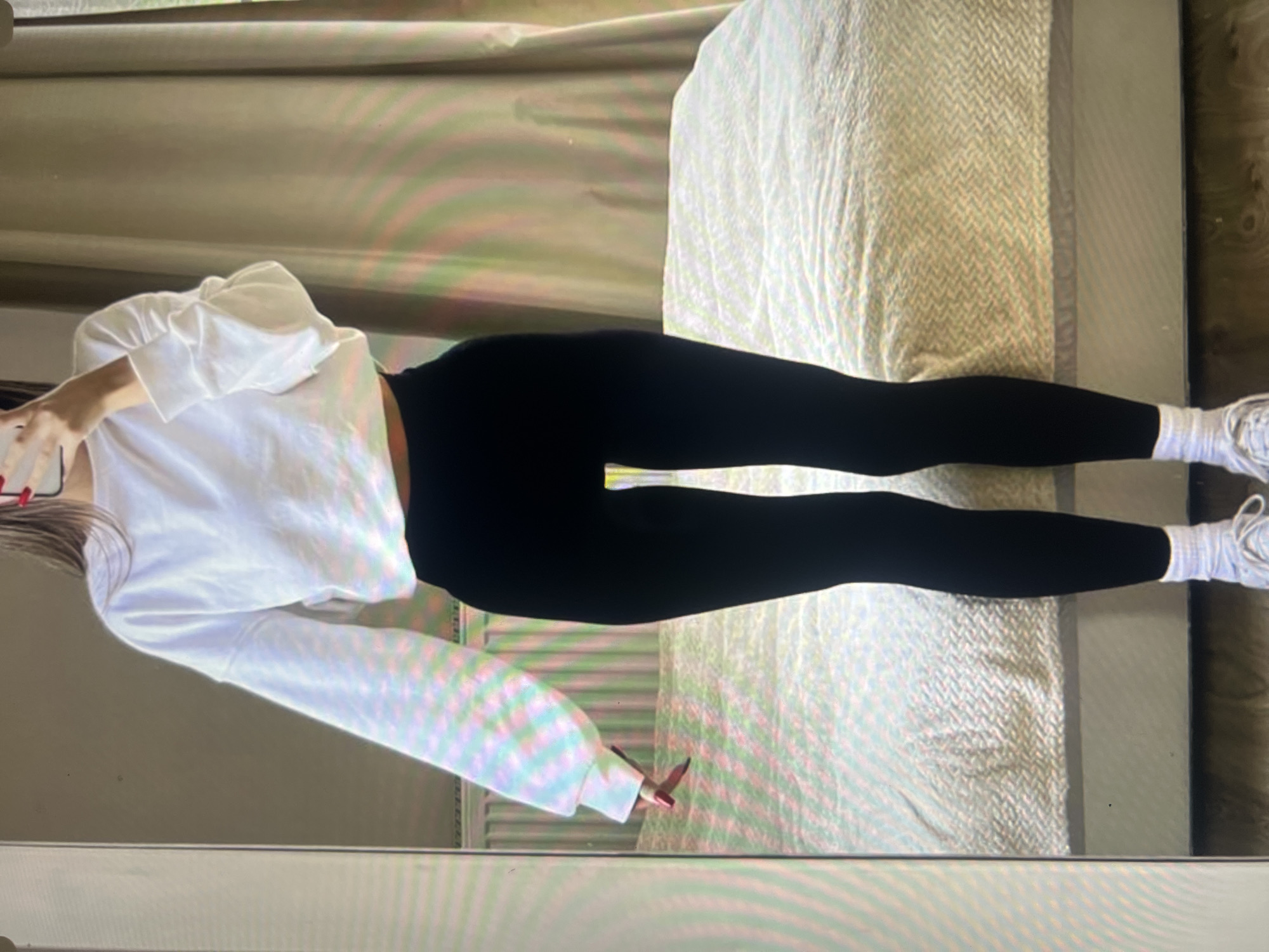 A person is standing in front of a mirror, wearing a white cropped sweatshirt, black leggings, white socks, and white sneakers. The person's hair is partially visible, and they are holding a phone in their hand. The background shows a bed with a beige and white patterned comforter and a beige curtain. The floor is made of wooden planks. The scene appears to be indoors in a bedroom.