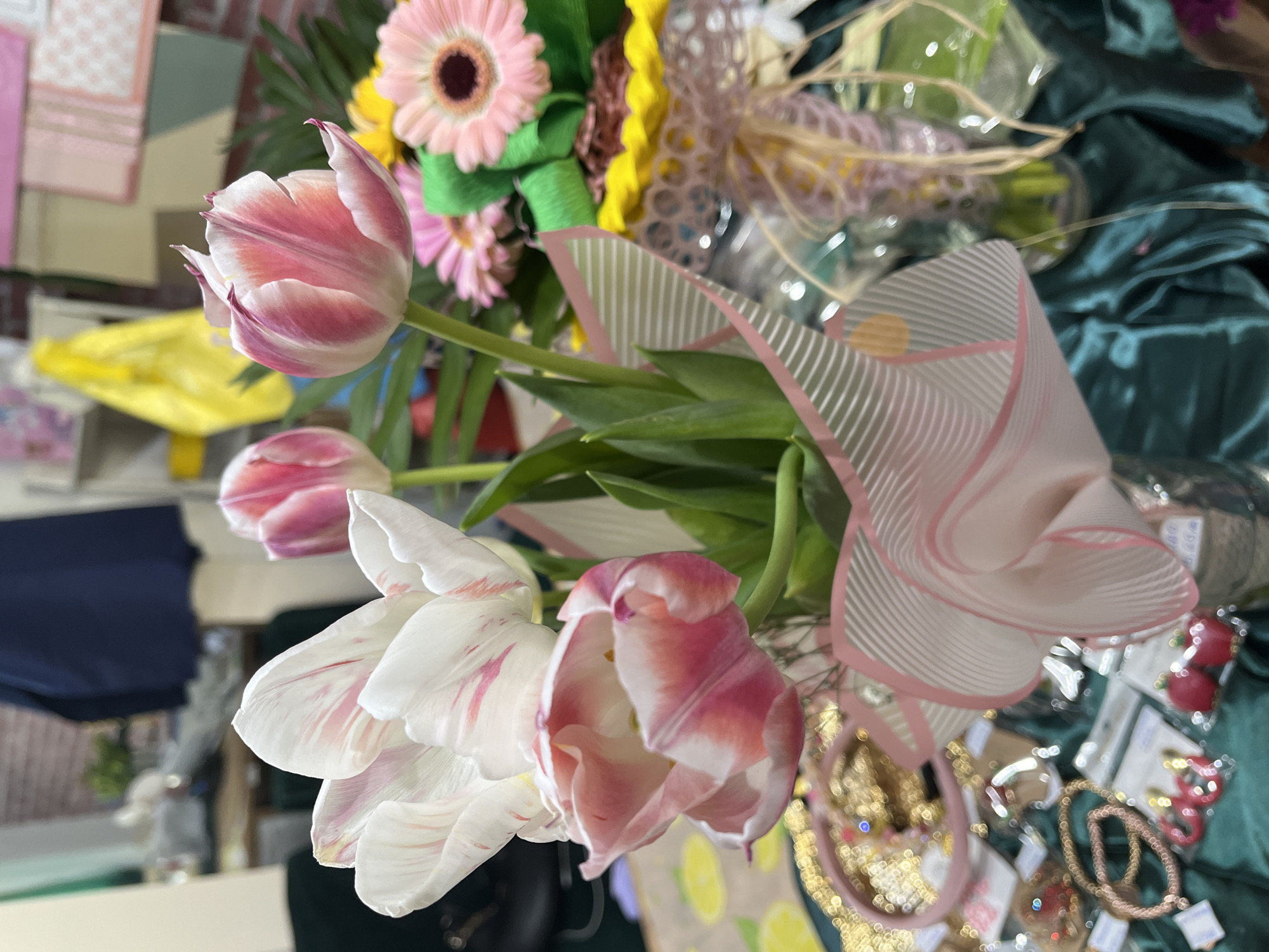 A close-up shot displays a bouquet of flowers dominated by white and pink tulips. A pink gerbera daisy and other floral elements are also visible. The arrangement is styled with decorative ribbon. In the background, items that appear to be jewelry and small decorative objects sit atop a draped teal fabric. The photograph was taken at a shop selling souvenirs in the city of Burgas, Bulgaria.
