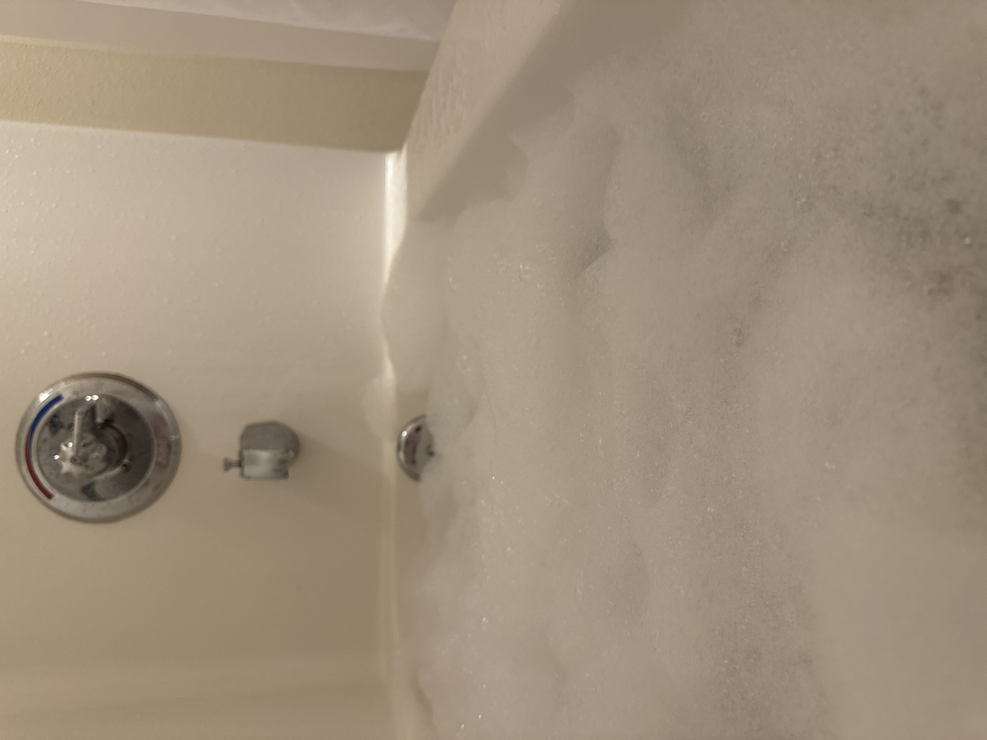 The image shows a close-up view of a bathtub filled with soap bubbles. The bathtub is white, and the bubbles obscure much of the scene. The bathtub fixtures, including the water temperature control and shower diverter, are visible on the left side of the frame. There is no evidence to determine the location in the city of Anchorage, United States of America. The scene includes a bathtub and soap bubbles.